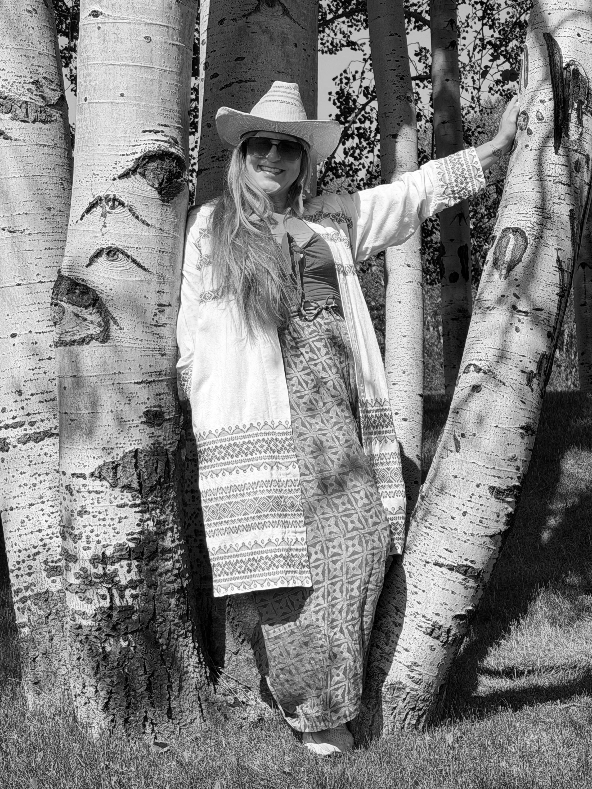 A woman in sunglasses and a cowboy hat standing among trees, smiling and leaning against a tree trunk, in a black and white photograph.