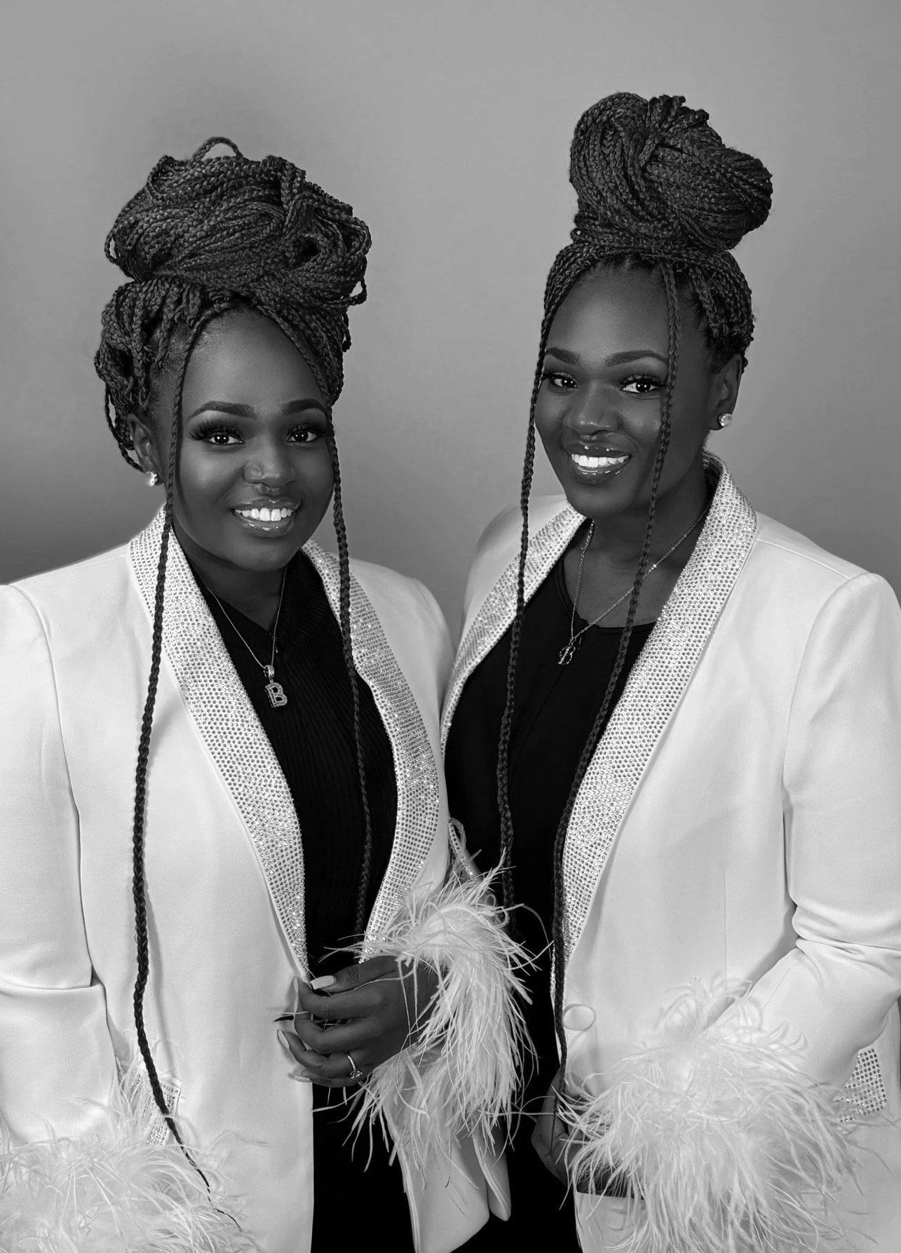 Two women smiling, dressed in white blazers with feather details, wearing similar hairstyles with braids styled in tall updos.