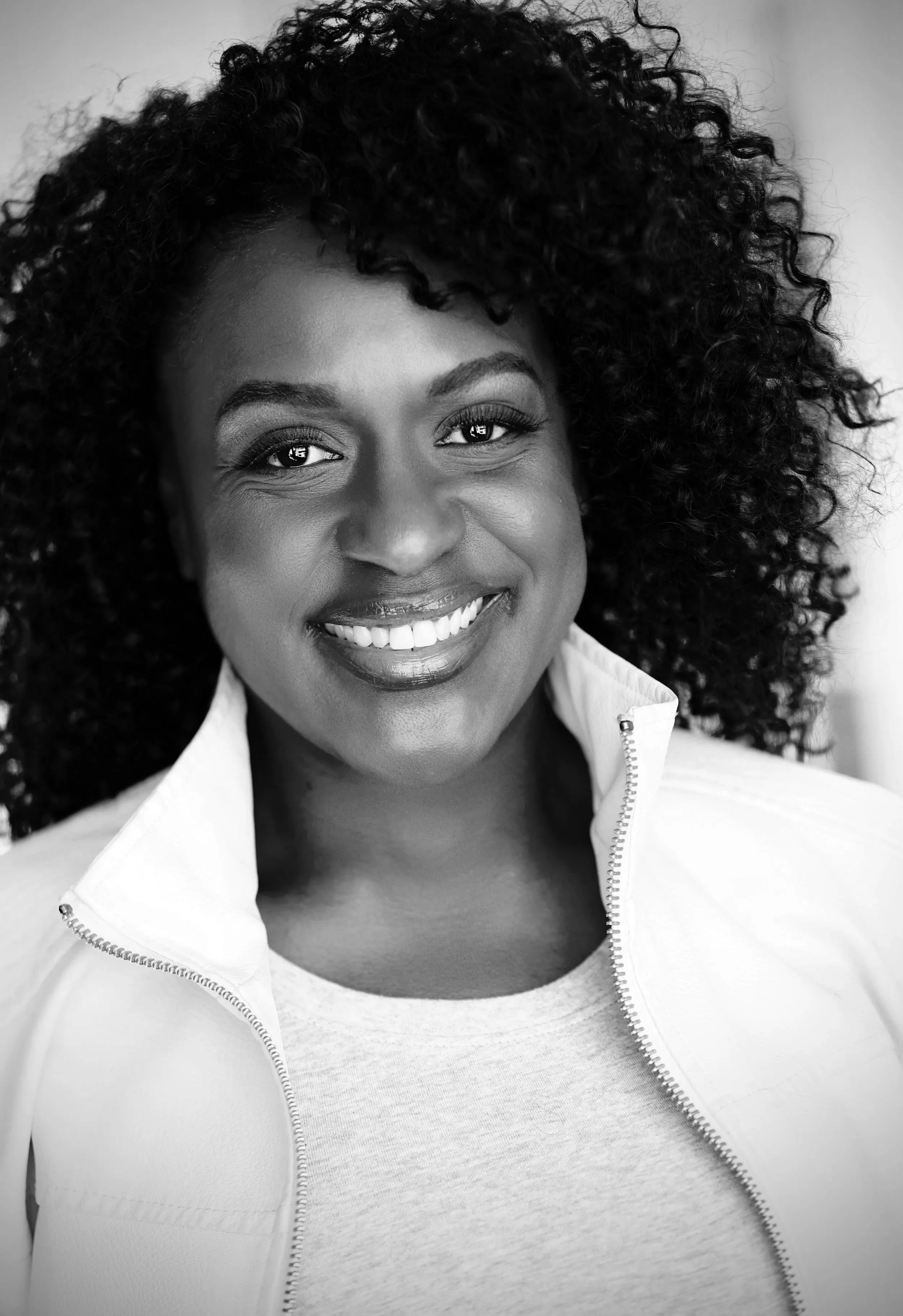 Close-up black and white photo of a woman smiling, with curly hair, wearing a zip-up jacket over a t-shirt.