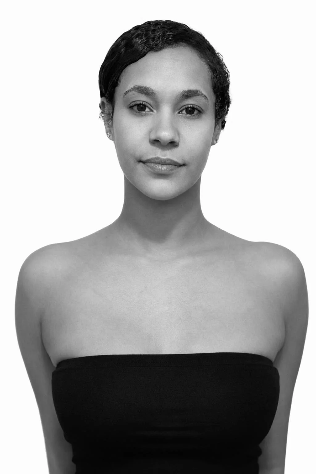 Black and white portrait of a young woman with short, curly hair, wearing a strapless black top, looking directly at the camera with a neutral expression.