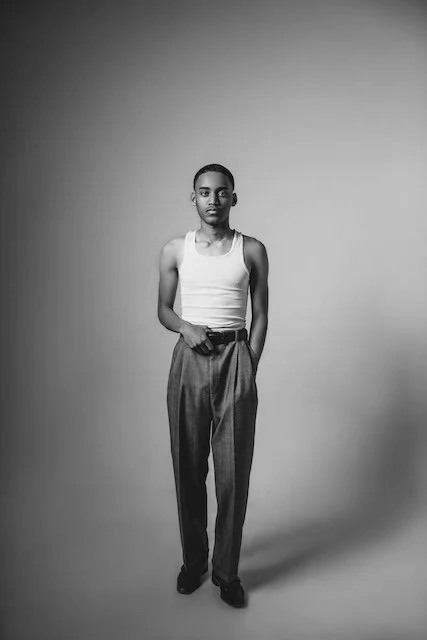Black and white photo of a man standing and facing forward, wearing a white tank top and high-waisted trousers.