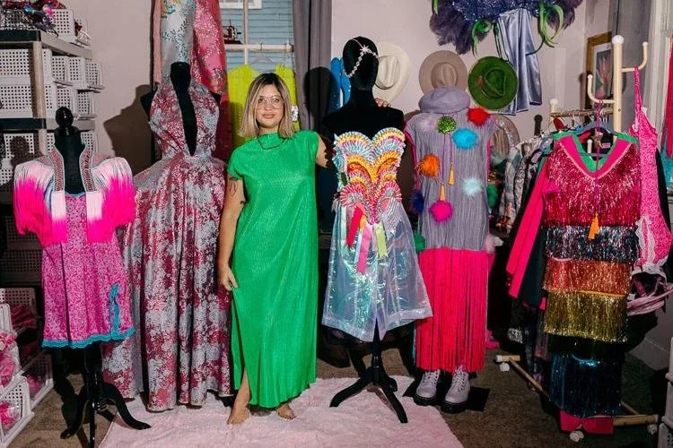 A woman in a bright green dress standing among colorful, eccentric costumes on dress forms in a costume shop.