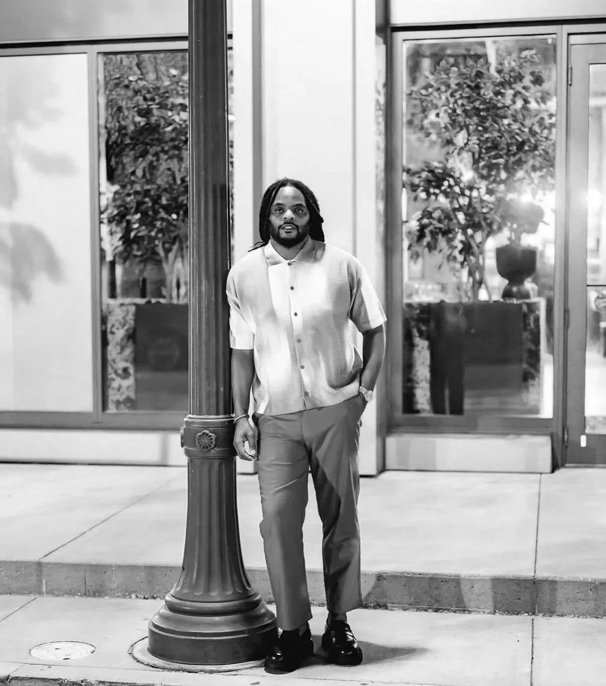A man with dreadlocks and a beard stands on a sidewalk at night, leaning against a streetlamp. He wears a short-sleeved, striped shirt and trousers, with his left hand in his pocket. Behind him are glass doors reflecting trees and potted plants, illuminated by lighting.