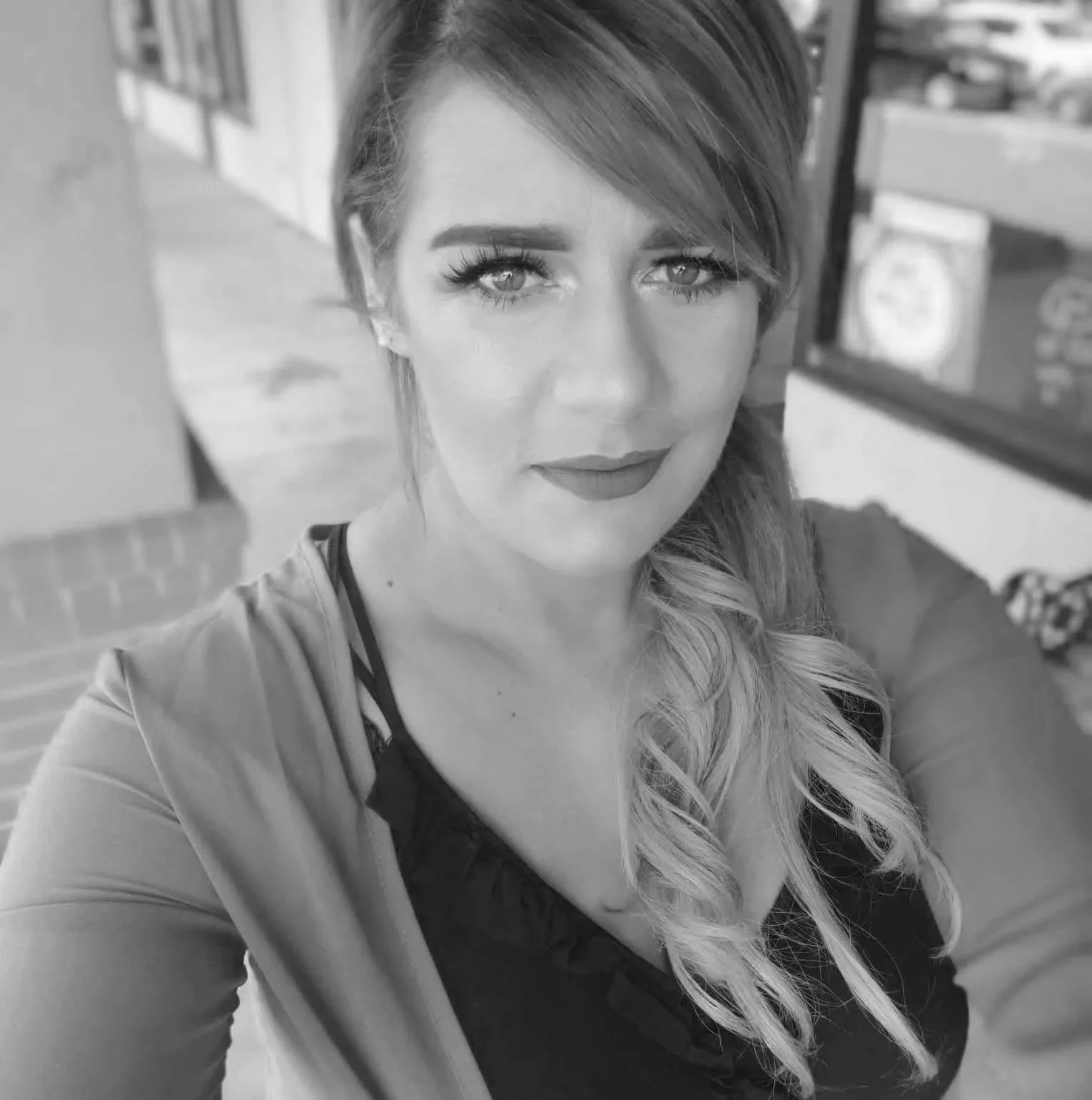 A woman with long, wavy hair and makeup, including eyeliner and lipstick, taking a selfie indoors with a blurred background that appears to be a store.