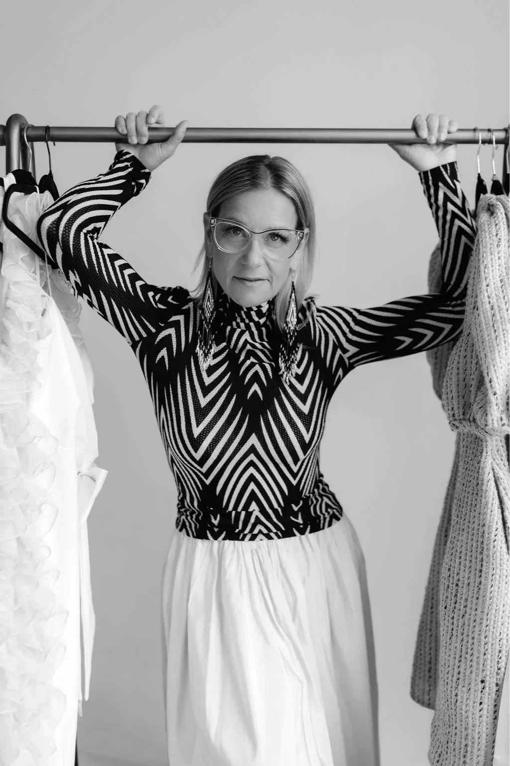 A woman wearing a zebra pattern long sleeve top, glasses, and earrings, standing behind a clothing rack with various garments, holding a metal rod above her head.
