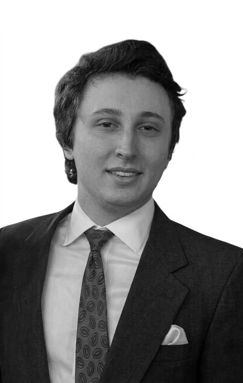 Black and white portrait of a young man in formal attire, wearing a suit, tie, and a pocket square, with short wavy hair and a slight smile.