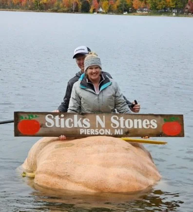 Two people floating in a pumpkin in a lake holding a sign that reads 'Sticks N Stones, Pierson, MI' with trees and fall foliage in the background.