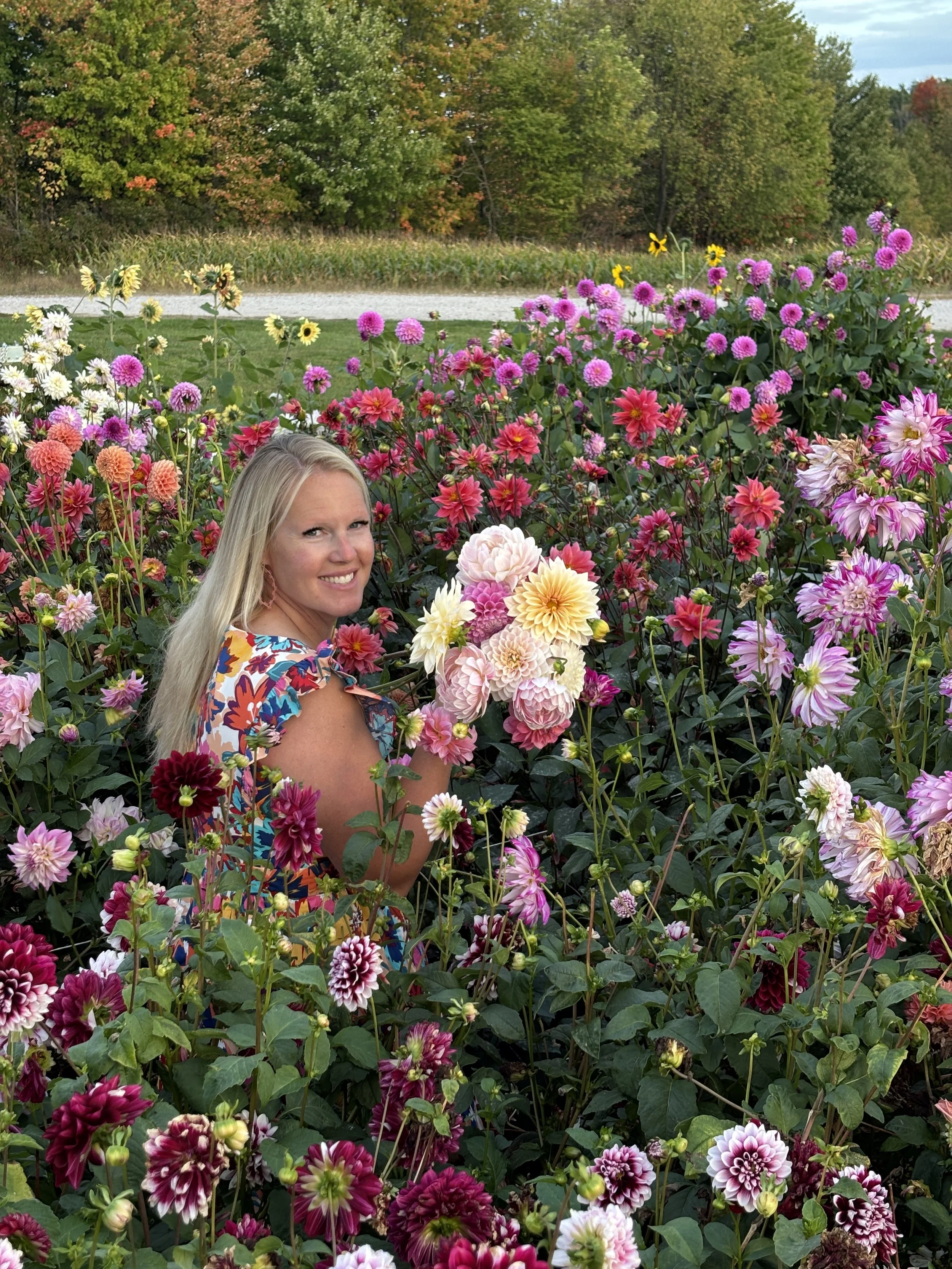 A smiling woman with long blonde hair in a colorful floral dress among a vibrant garden of pink, purple, white, and red flowers with green foliage, during daytime.
