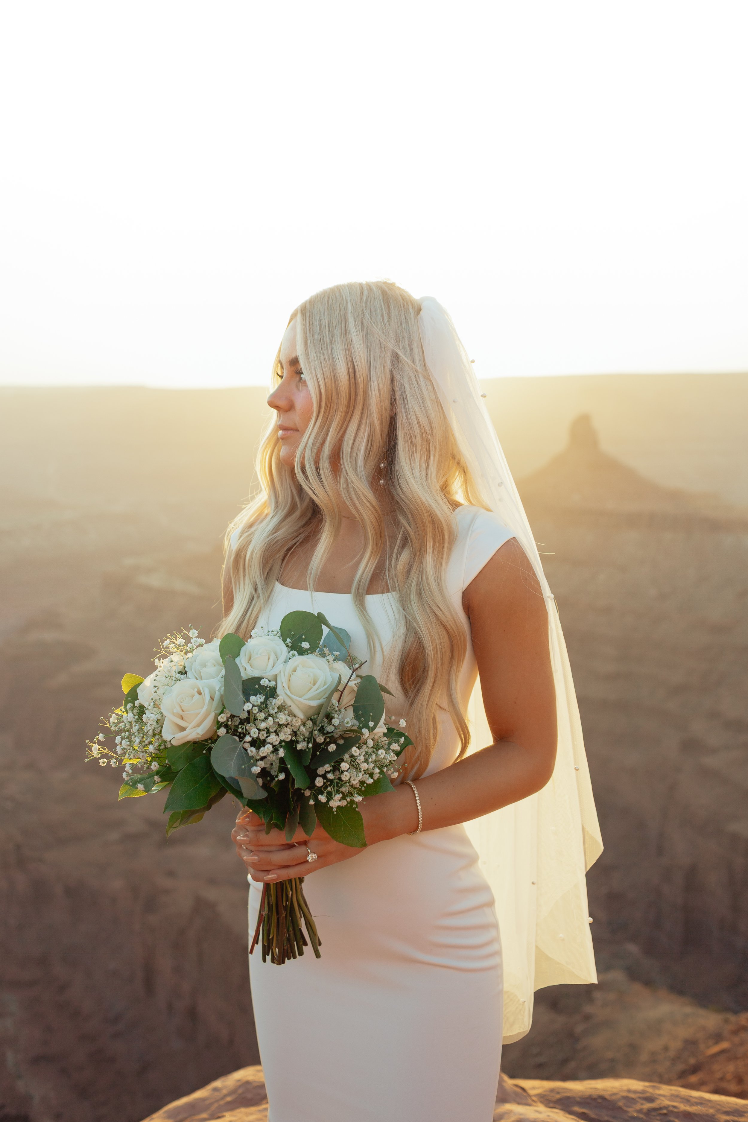 Bride in a white wedding dress holding a bouquet of white roses and greenery, standing outdoors at sunset with a canyon landscape in the background at Dead Horse Point State Park in Moab, Utah.