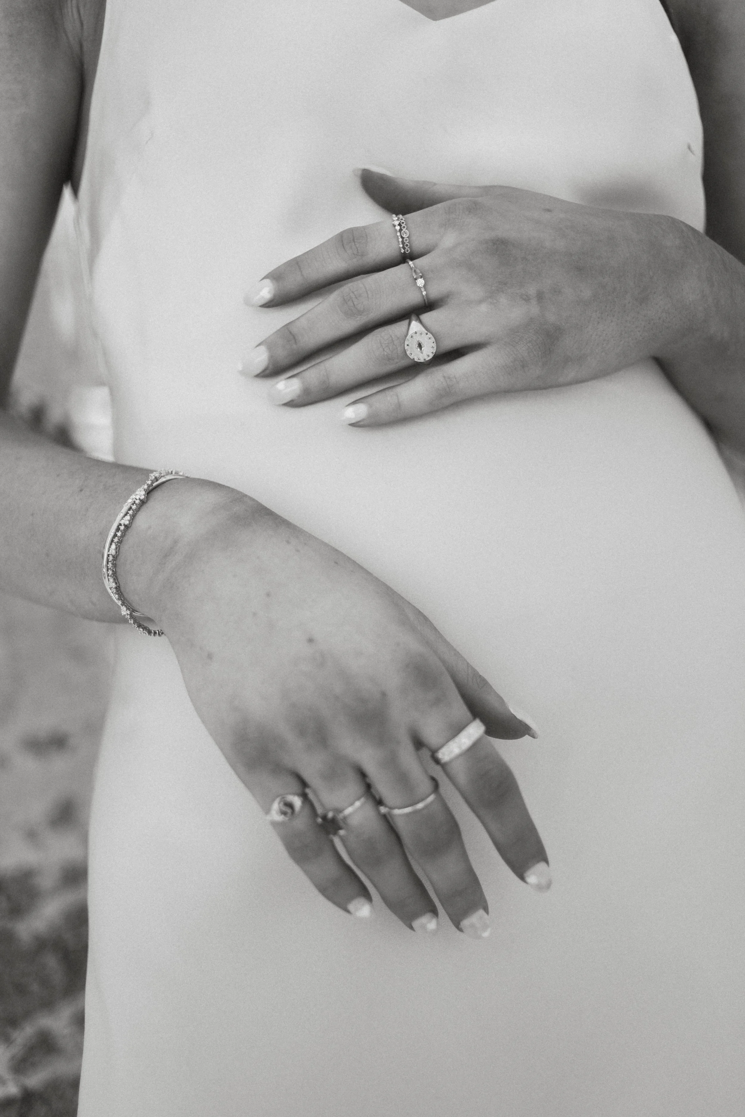 Close-up of a woman's hands and part of her arm, adorned with rings and bracelets, resting on her abdomen, which is covered by a smooth, light-colored fabric. Taken in Deer Valley / Park City Utah.