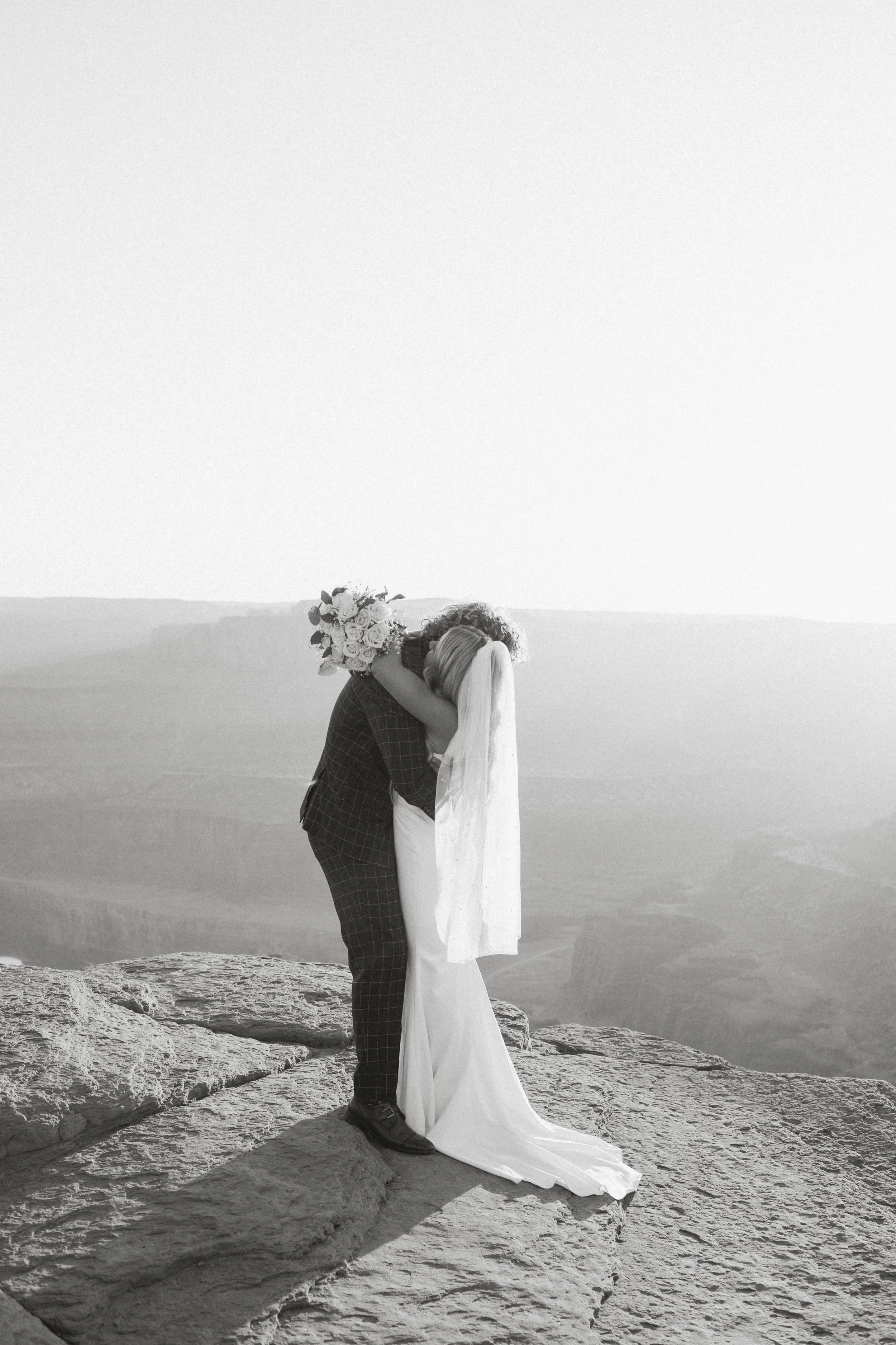 Black and white photo of a bride and groom embracing on a rocky cliff with expansive canyon views in the background at Dead Horse Point State Park in Moab, Utah.