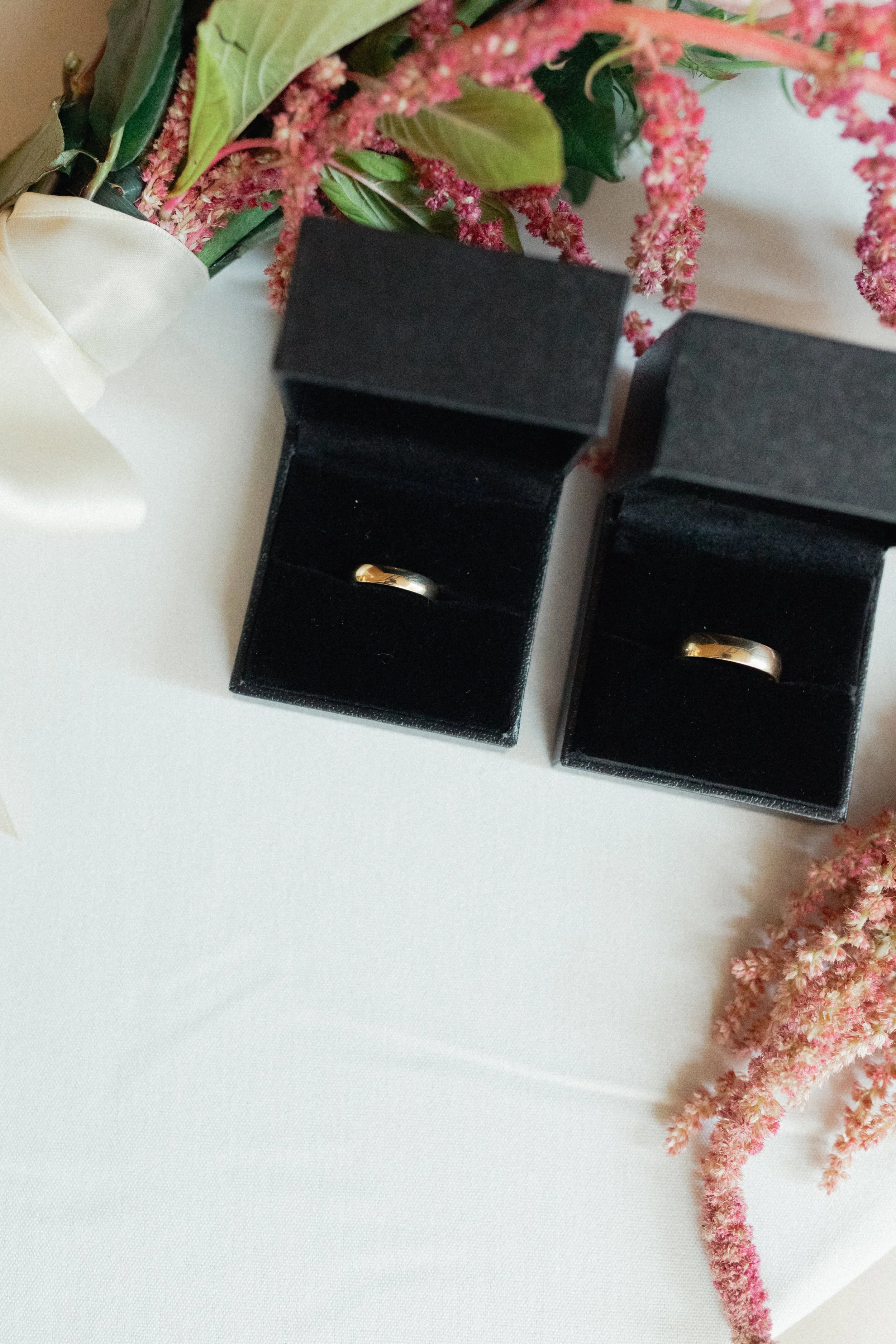 Two black jewelry boxes each containing a gold ring, surrounded by pink and green flowers on a white surface. Taken at Caldera House.