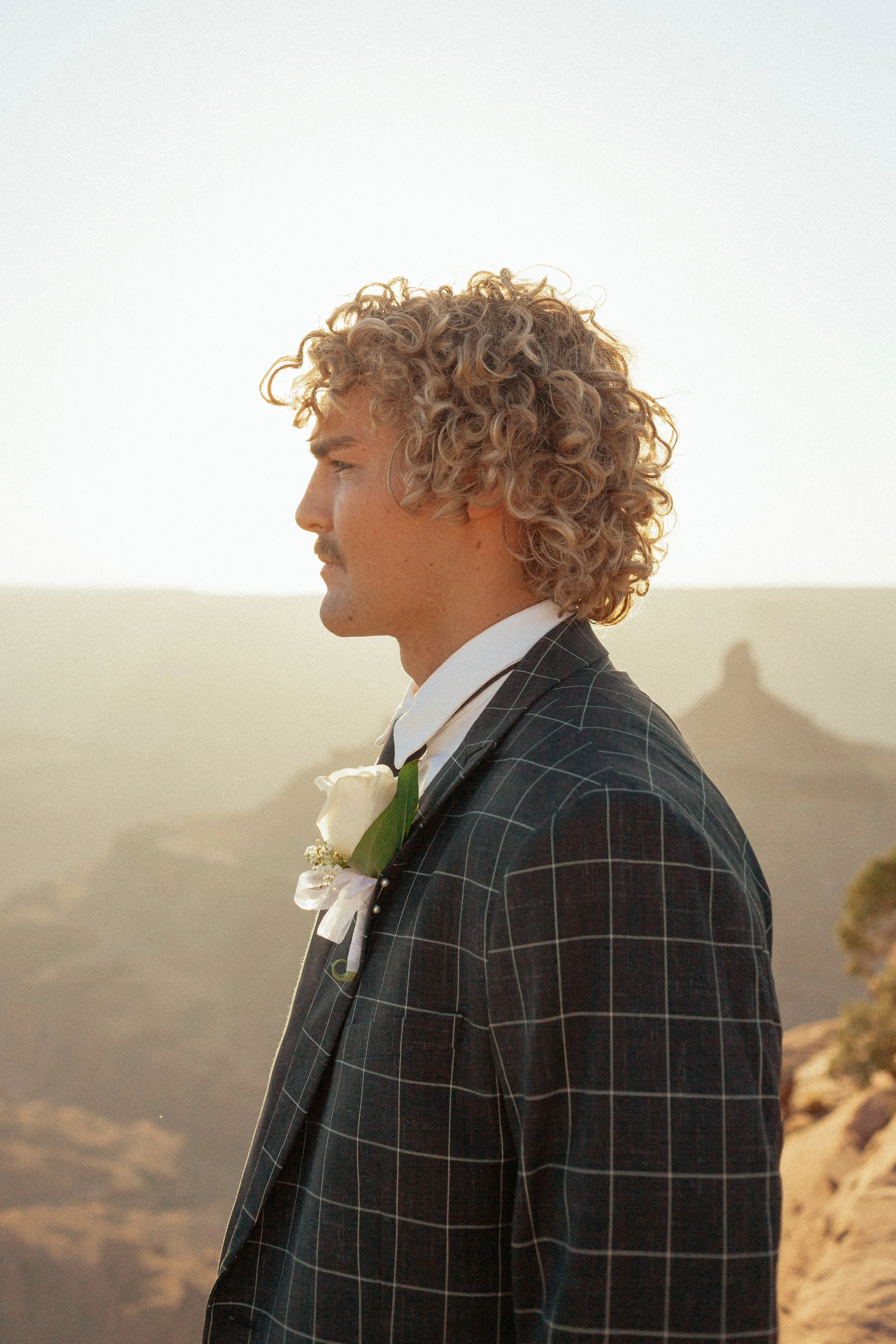 A man with curly blonde hair is standing in profile, outdoors, wearing a checkered suit with a white shirt and a boutonnière. The background features a hazy landscape with hills and a rock formation. at Dead Horse Point State Park in Moab, Utah.
