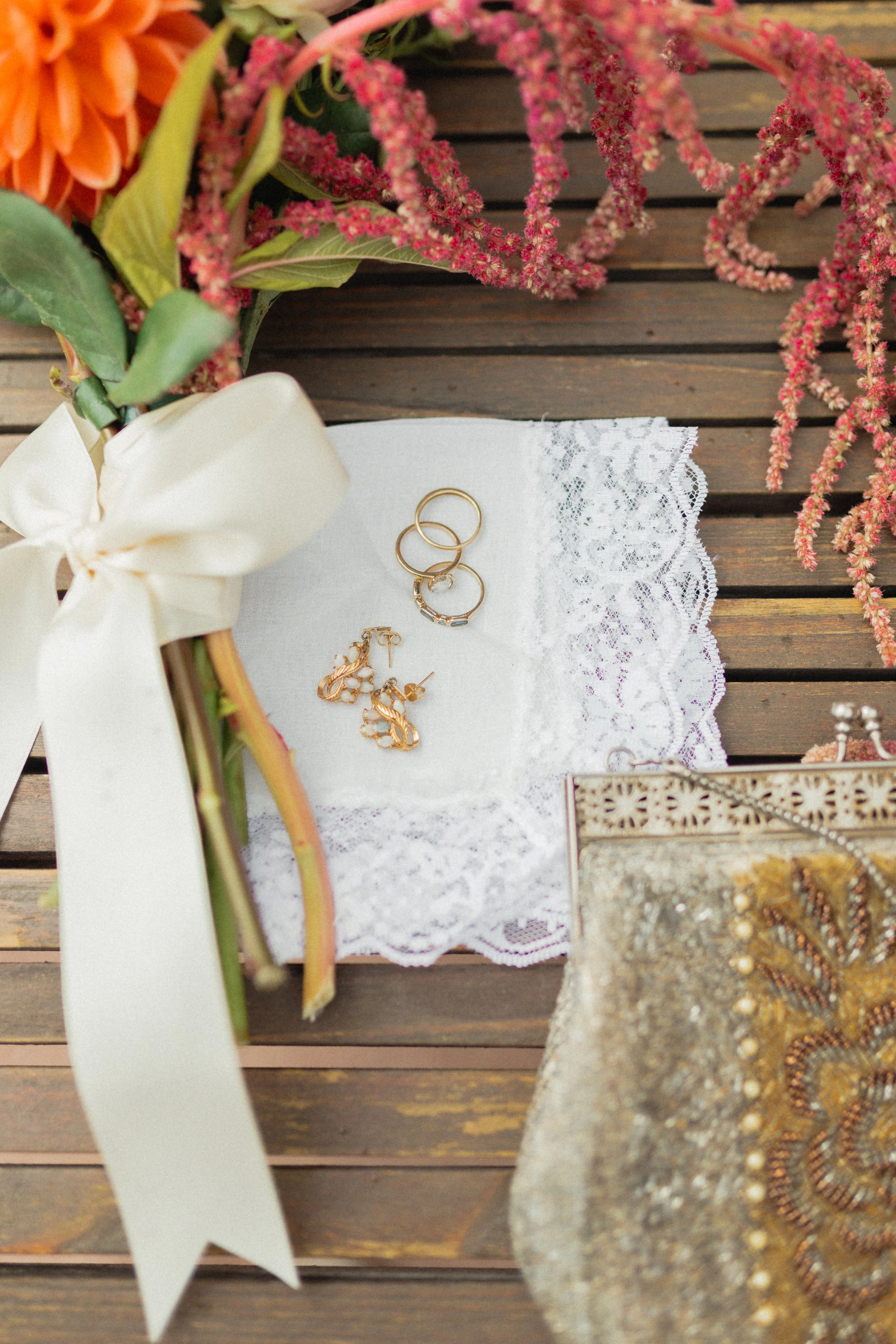 A wedding bouquet with orange and pink flowers, a white ribbon, gold rings and jewelry on a white lace cloth, and a gold and beaded purse, all on a wooden surface. Bridal details in Teton Village, Jackson Hole, Wyoming.