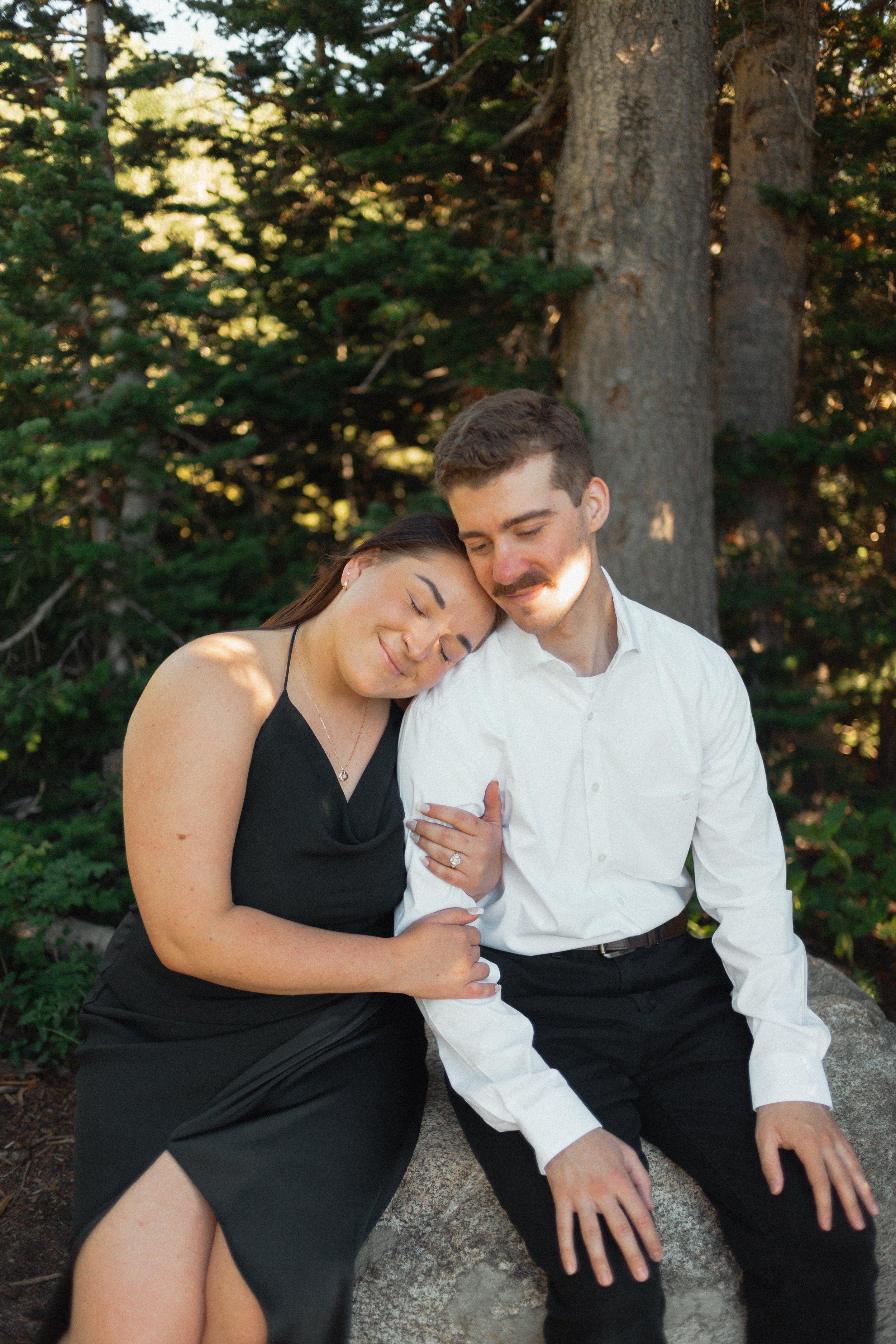 A couple sitting on a rock in a wooded area, with the woman resting her head on the man's shoulder, both appearing content and affectionate. Taken at Albion Basin, in Alta Utah.