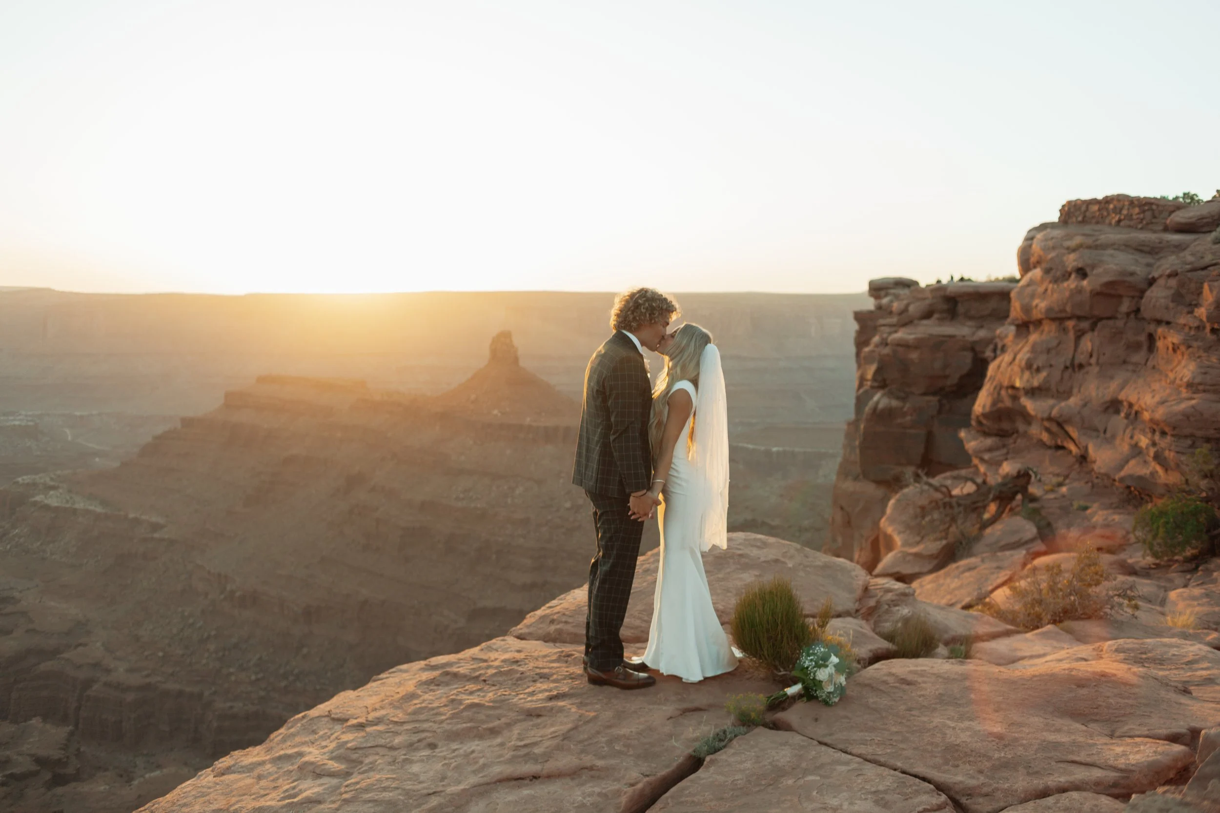 A couple in wedding attire sharing a kiss on a rocky cliff at sunset, with a bouquet of flowers on the ground nearby, at Dead Horse Point State Park in Moab, Utah.