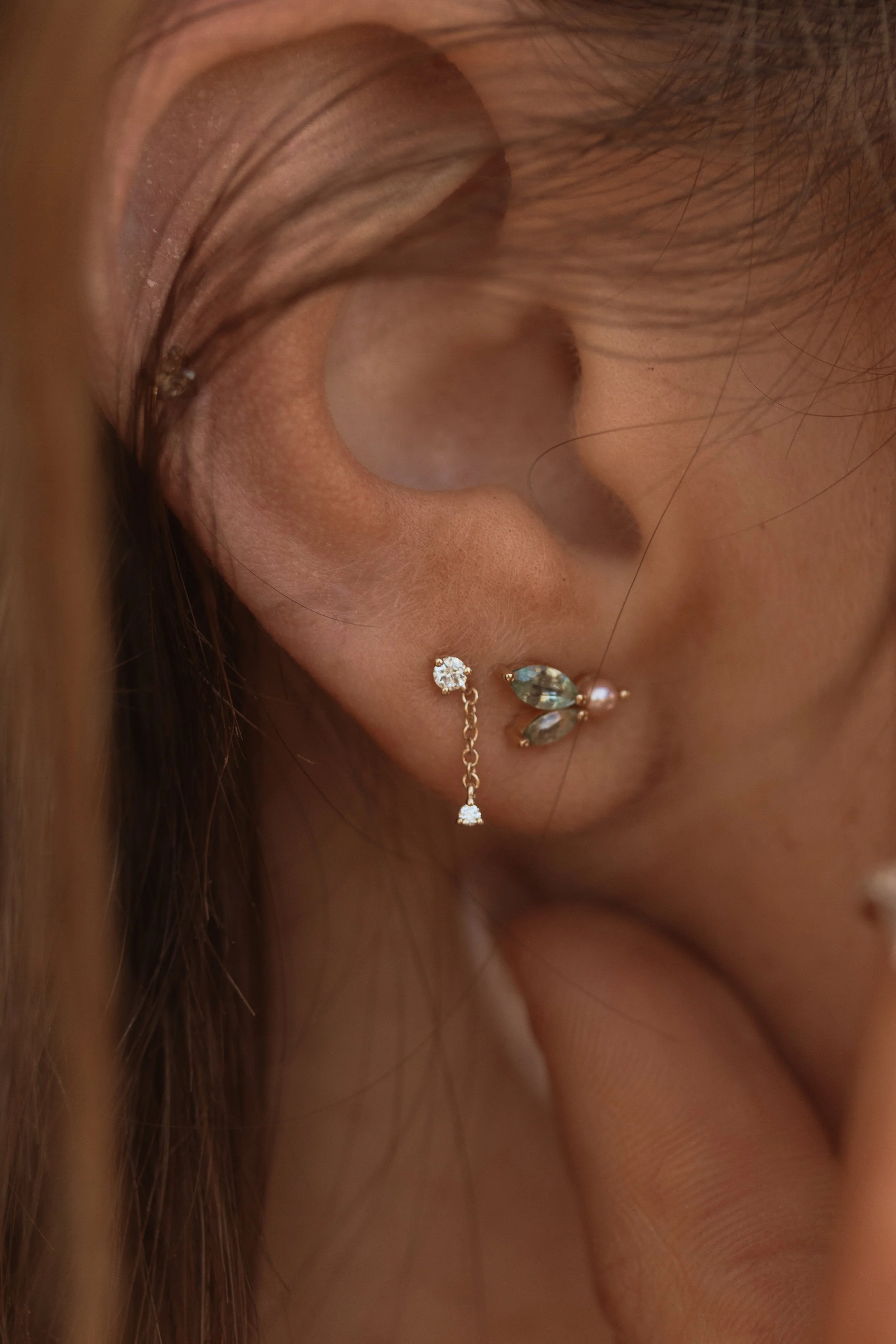 Close-up of a woman's ear with three earrings: a small diamond stud, a chain with a small diamond, and a pearl with a butterfly-shaped crystal. Shot for Park City based fine jewelry brand.