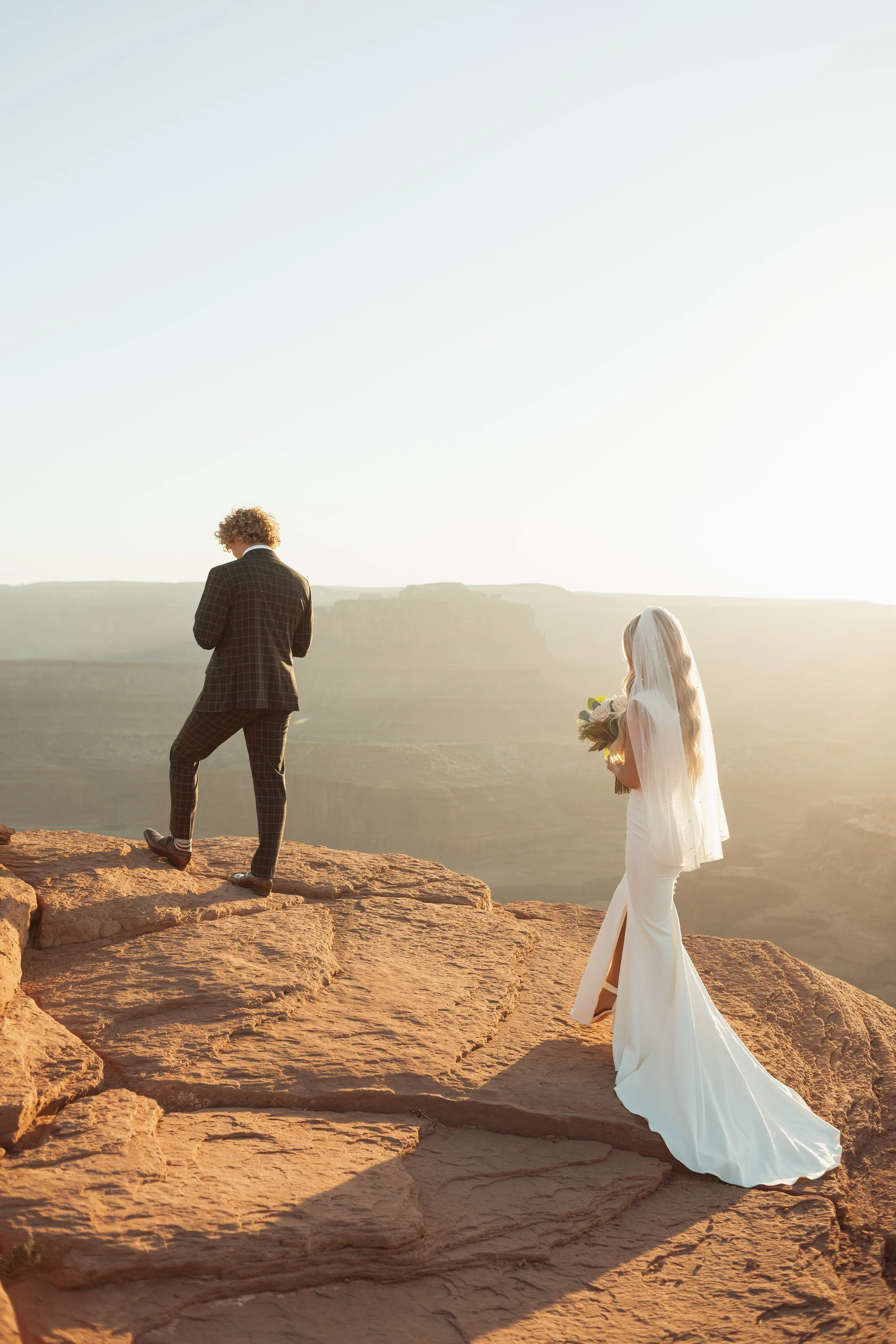A bride in a white wedding dress and veil holding a bouquet standing on rocks, facing a groom in a suit, on a scenic desert overlook at sunset at Dead Horse Point State Park in Moab, Utah.