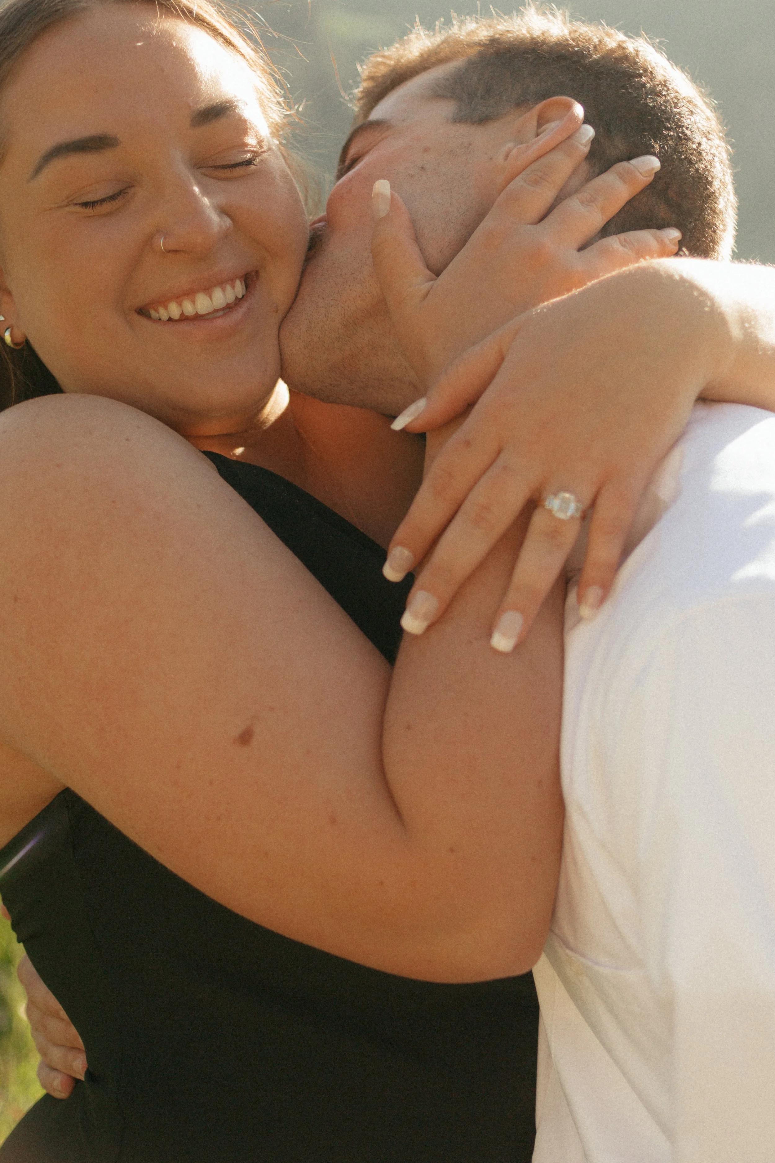 Engagement photos of smiling woman with a nose ring and a ring on her finger embracing and kissing a man outdoors with sunlight at Albion Basin.
