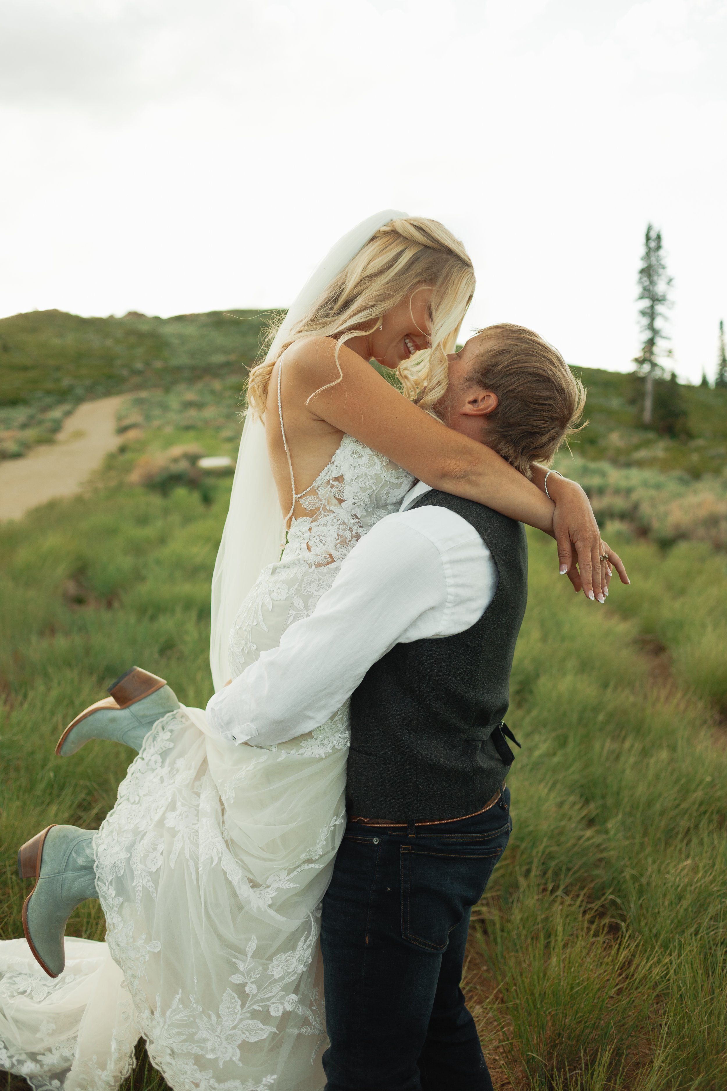 A happy bride in a white lace wedding dress and a groom in a white shirt and dark vest, outdoors in a grassy field, with the groom lifting the bride in his arms as they smile and embrace in Park City / Deer Valley Utah.