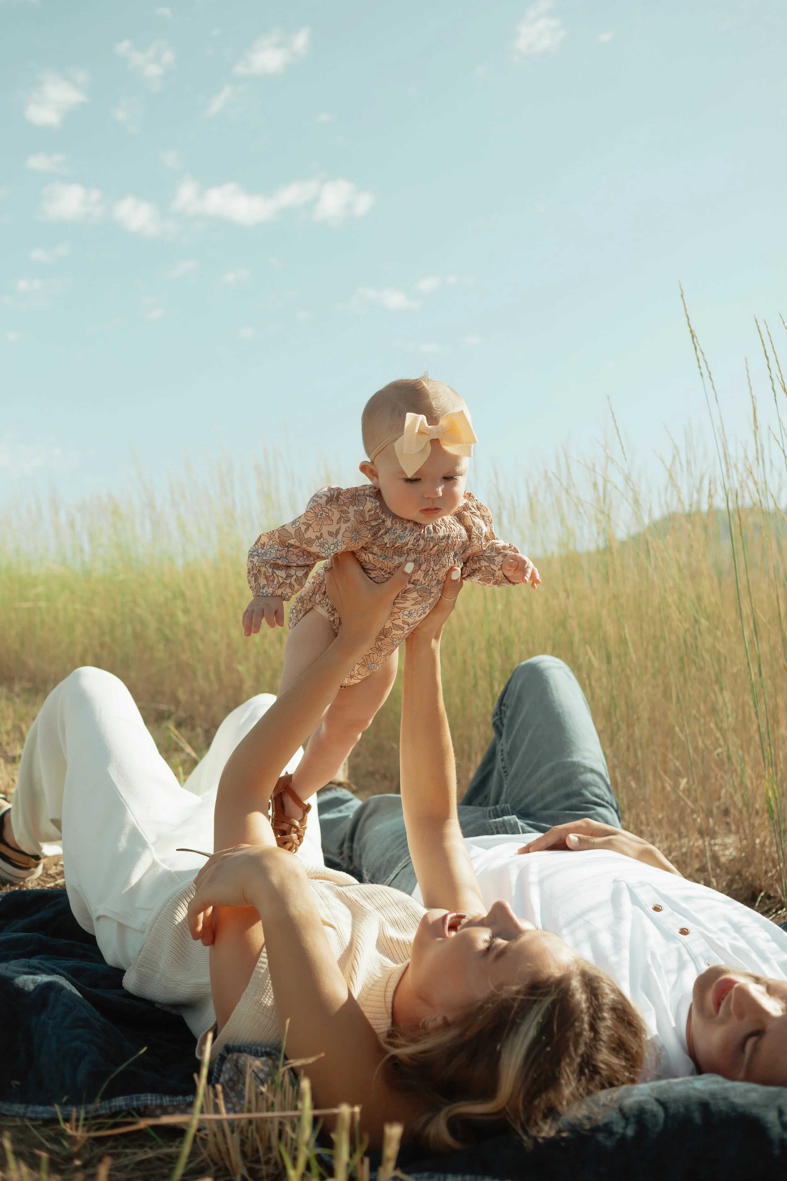 A woman and a man lying on a black blanket in a grassy field, playing with a baby girl who is being held up in the air. The scene is outdoors under a blue sky with some clouds.