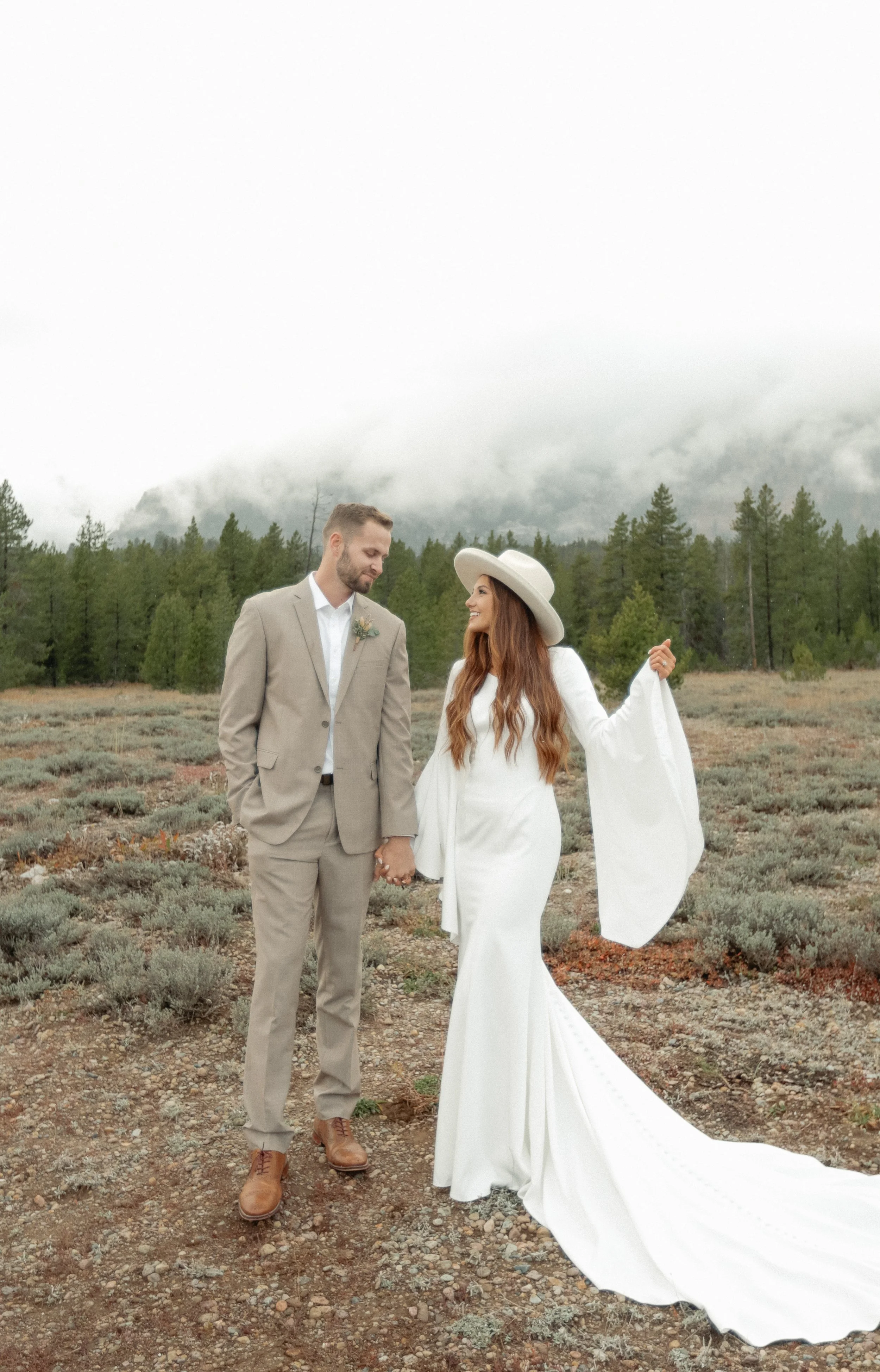 A bride and groom couple dressed in wedding attire holding hands in a natural outdoor setting with trees and cloudy sky in the background in Grand Teton National Park near Jackson Hole Wyoming.