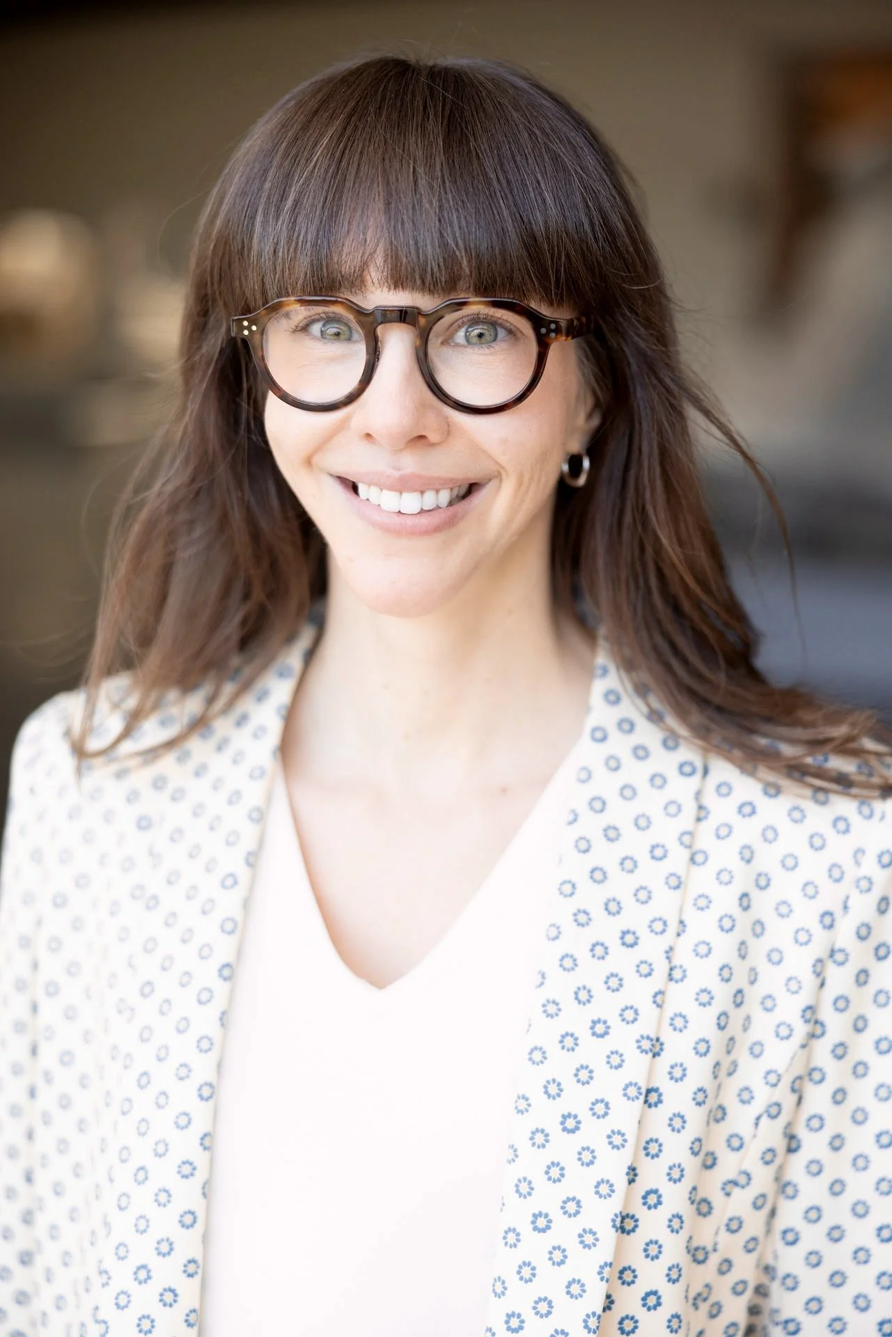 A woman, Ellen Kaross, with brown hair, glasses, and hoop earrings smiles outdoors, wearing a white top and a yellow blazer with blue polka dots.