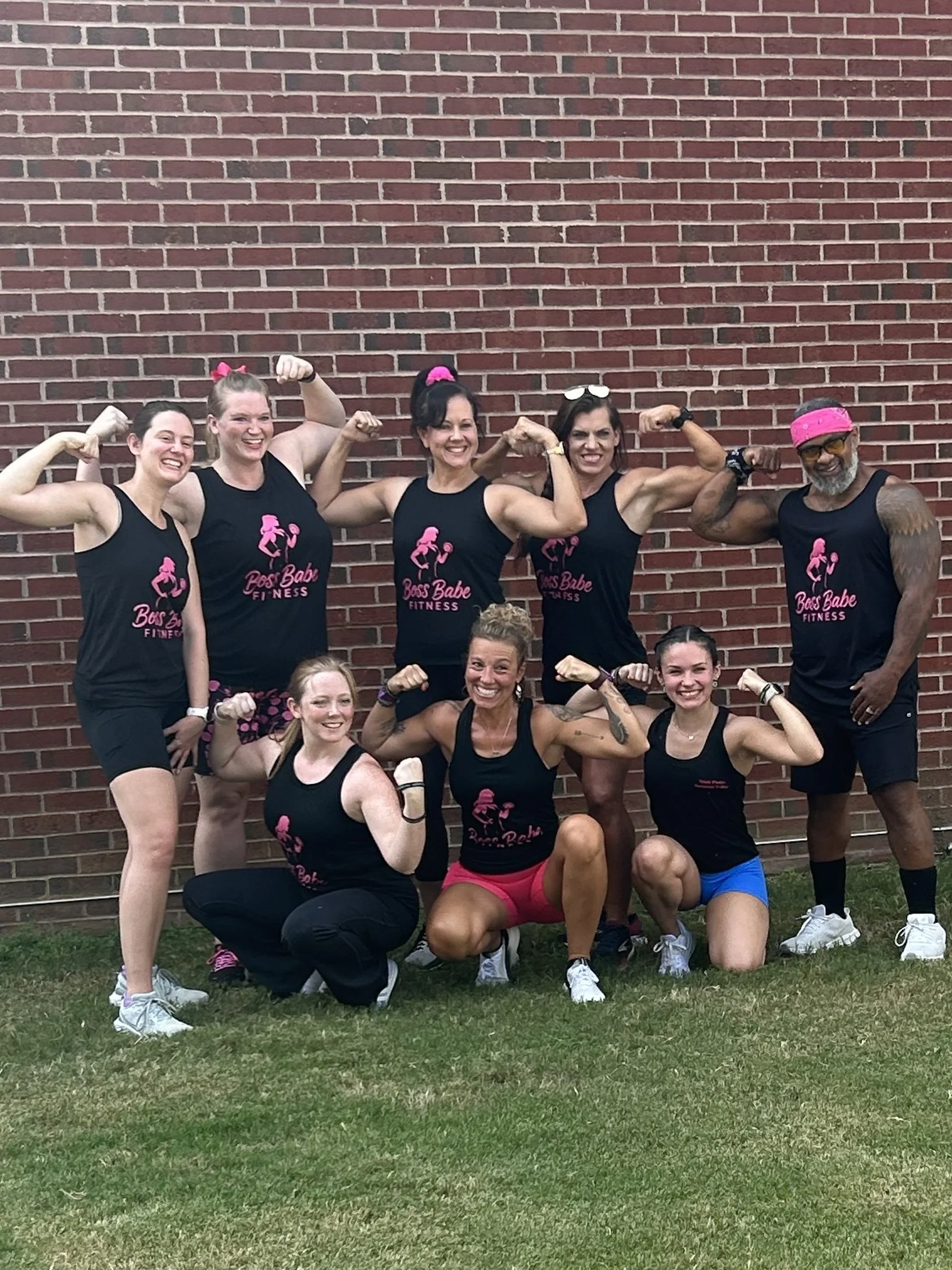 Group of women and one man wearing black fitness tank tops with pink text and graphics, posing in front of a brick wall. They are flexing their biceps and smiling.