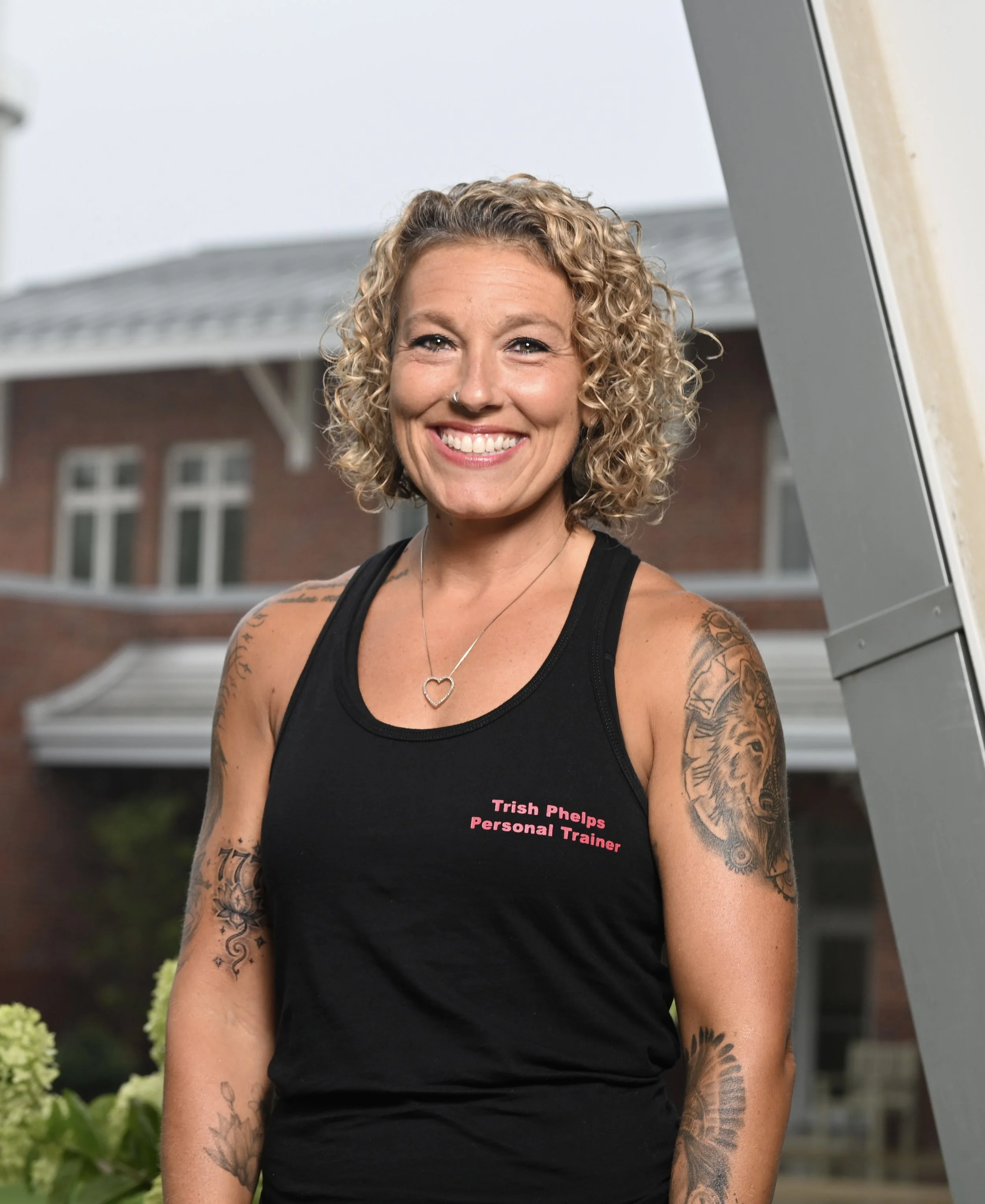 A smiling woman with curly blonde hair standing outdoors, wearing a black tank top with pink text that reads 'Trish Phelps Personal Trainer', tattoos on both arms, and a heart-shaped necklace.