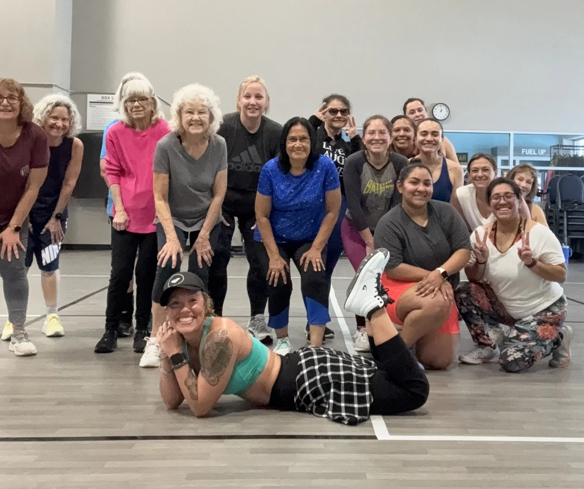 Group of women in a gym, some standing and some kneeling or lying down, smiling and posing for the camera.