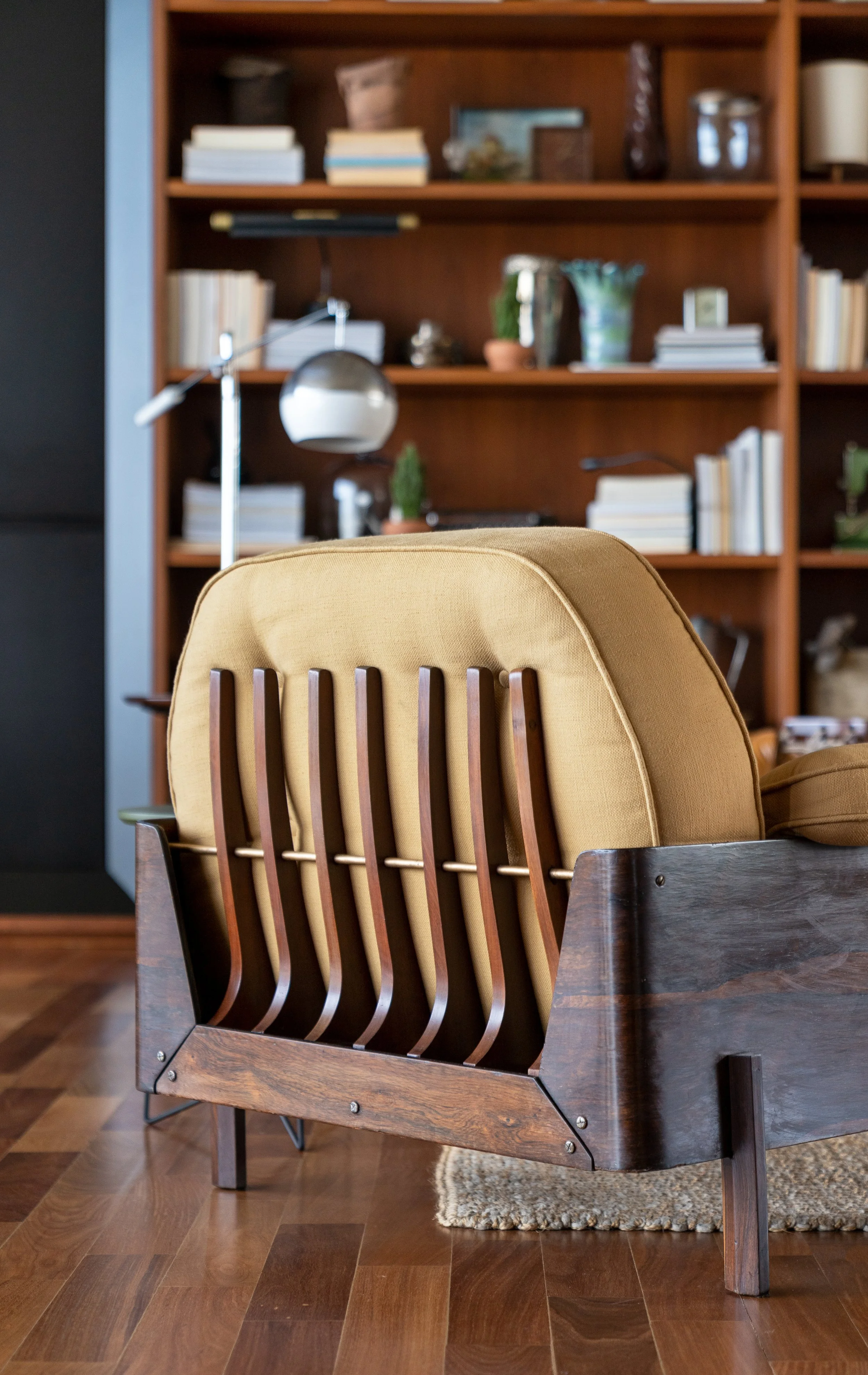 A mid-century modern armchair with a wooden frame and yellow cushion in front of a wooden bookshelf filled with books, plants, and decorative items, on a hardwood floor.