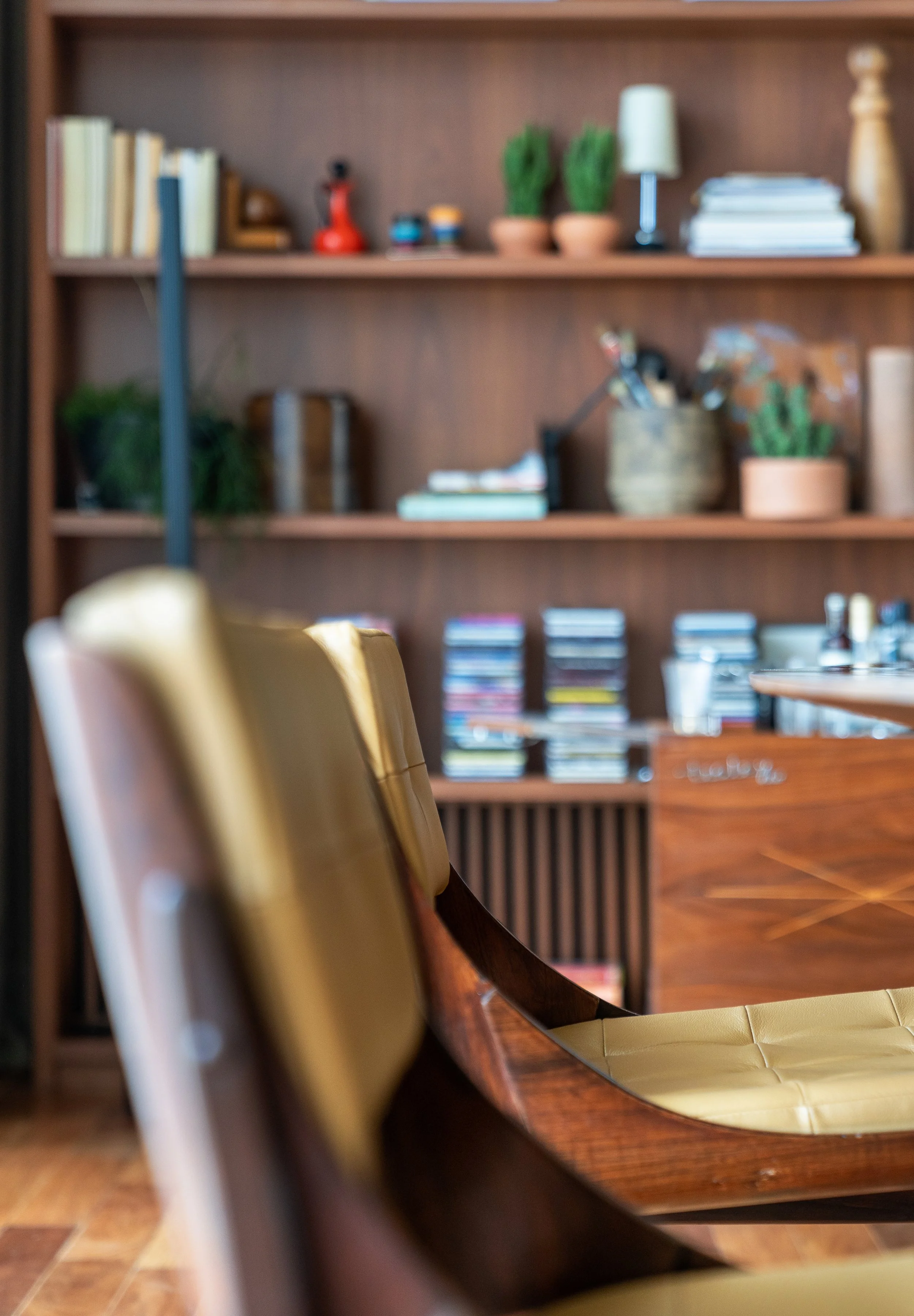 Close-up of a bookshelf with various books, plants, and decorative items, with a wooden chair with leather upholstery in the foreground.