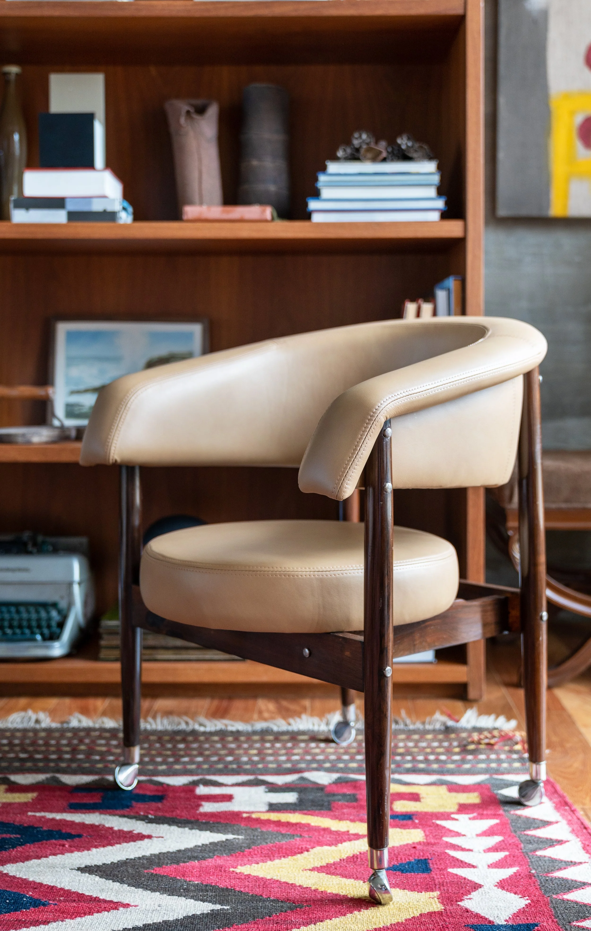 A beige vintage armchair with wooden frame and caster wheels, placed on a colorful geometric patterned rug in front of a wooden bookshelf containing various books, a framed photo, and decorative objects.