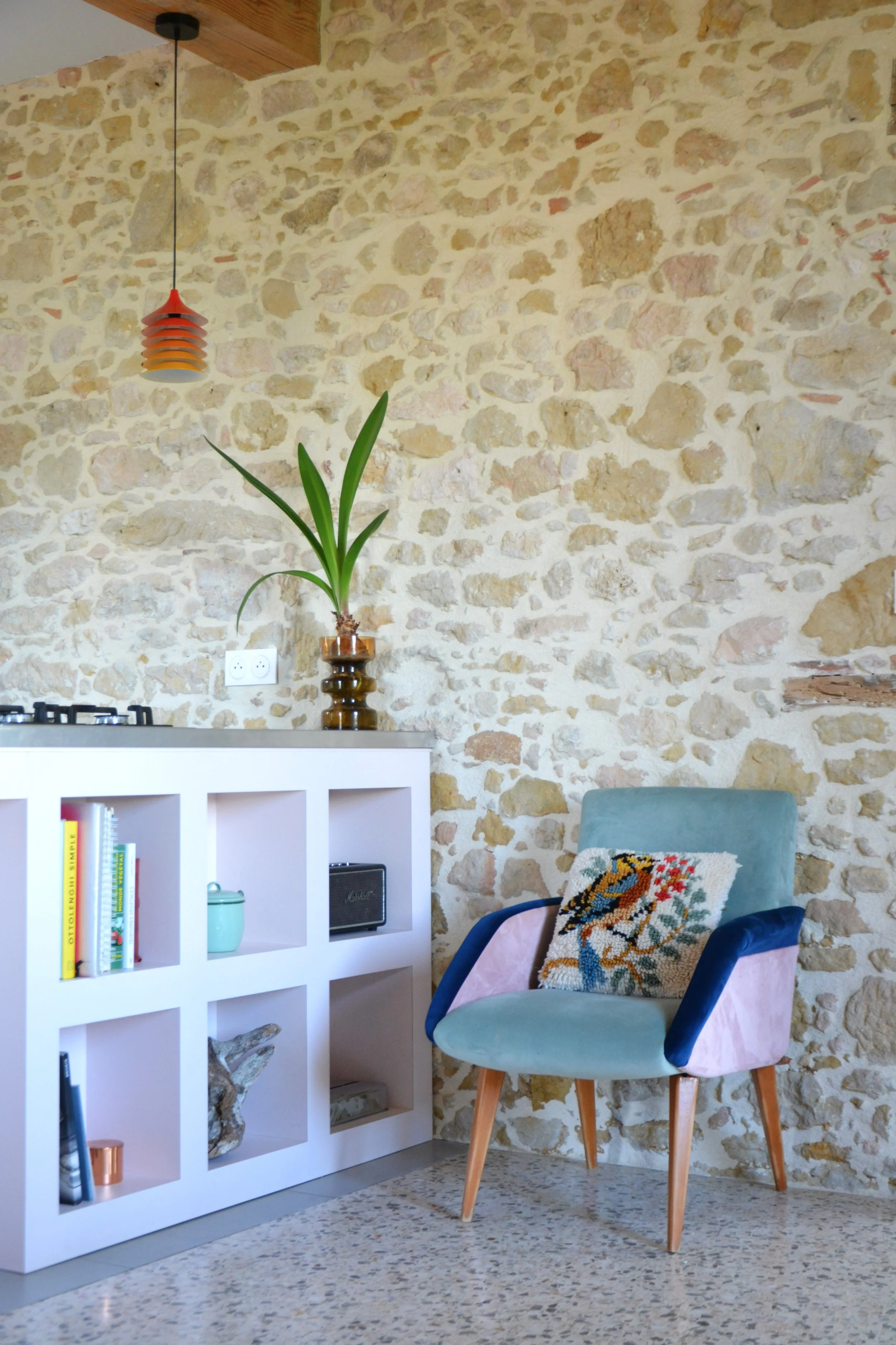 Cuisine sur-mesure avec un fauteuil vintage, des livres et objets décoratifs, un mur en pierre clair, et une suspension IKEA vintage orange et jaune.