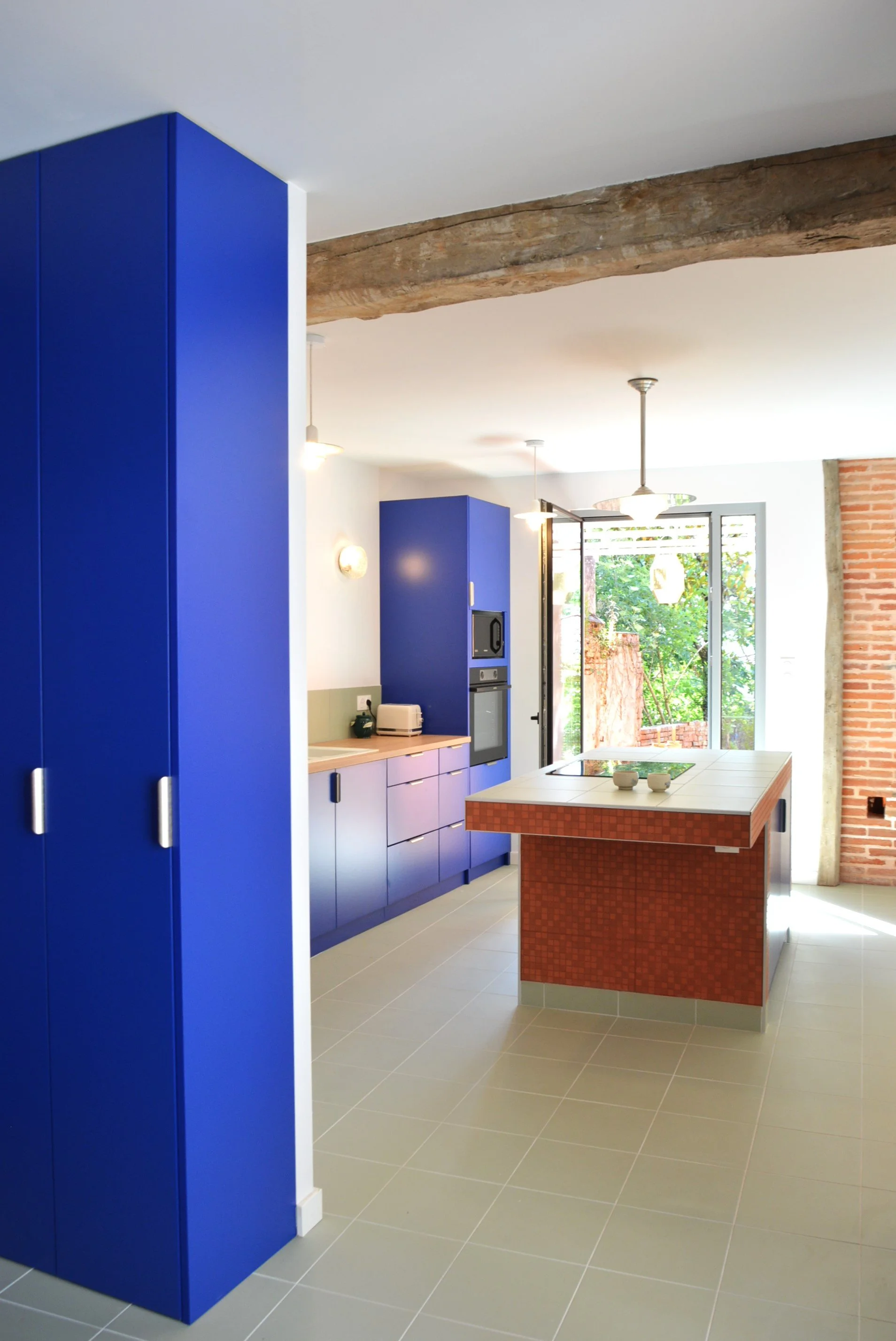 Cuisine moderne sur mesure, un îlot en terre cuite rouge, grand mur en brique, grand fenêtre avec vue sur un jardin verdoyant.