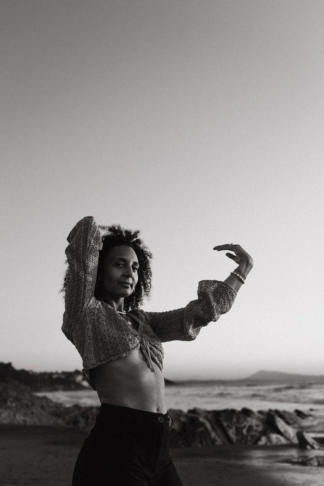 Une femme pose sur la plage pendant le coucher du soleil, portant un haut en crochet et un pantalon noir, dans une pose artistique avec un arrière-plan de ciel nuageux et de la mer.