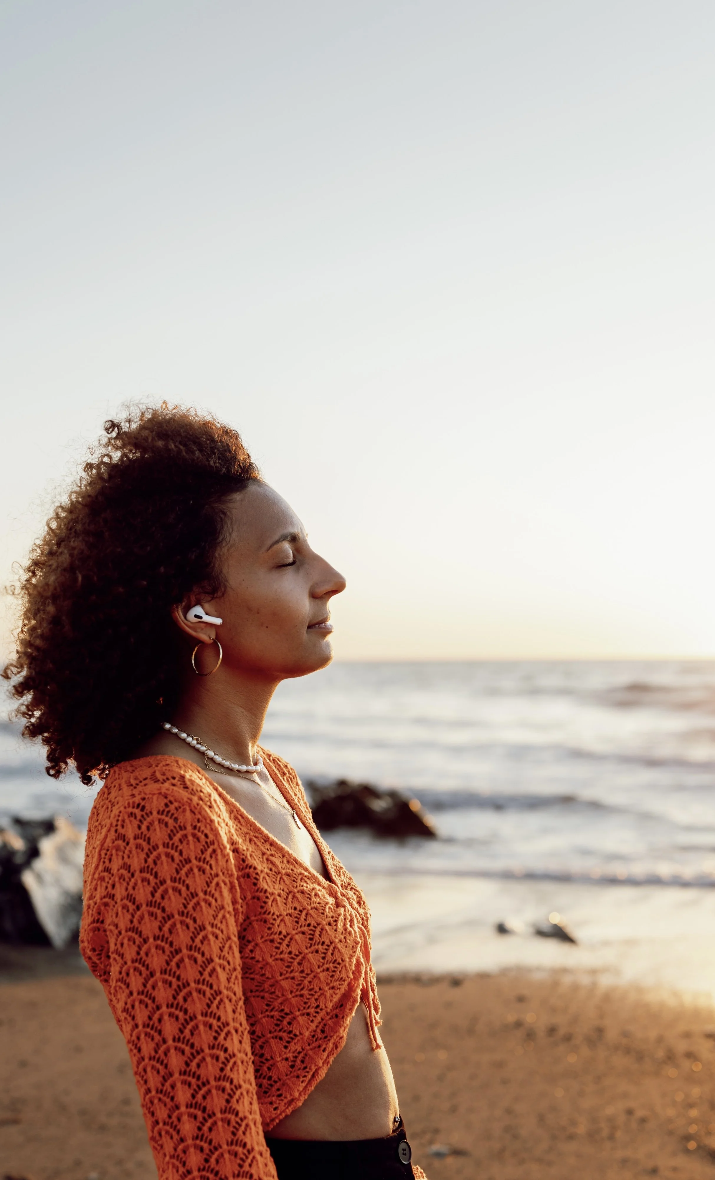 Une femme avec des cheveux bouclés, portant une chemise en crochet orange, regarde la mer au coucher du soleil, portant des écouteurs sans fil et des bijoux, sur une plage.