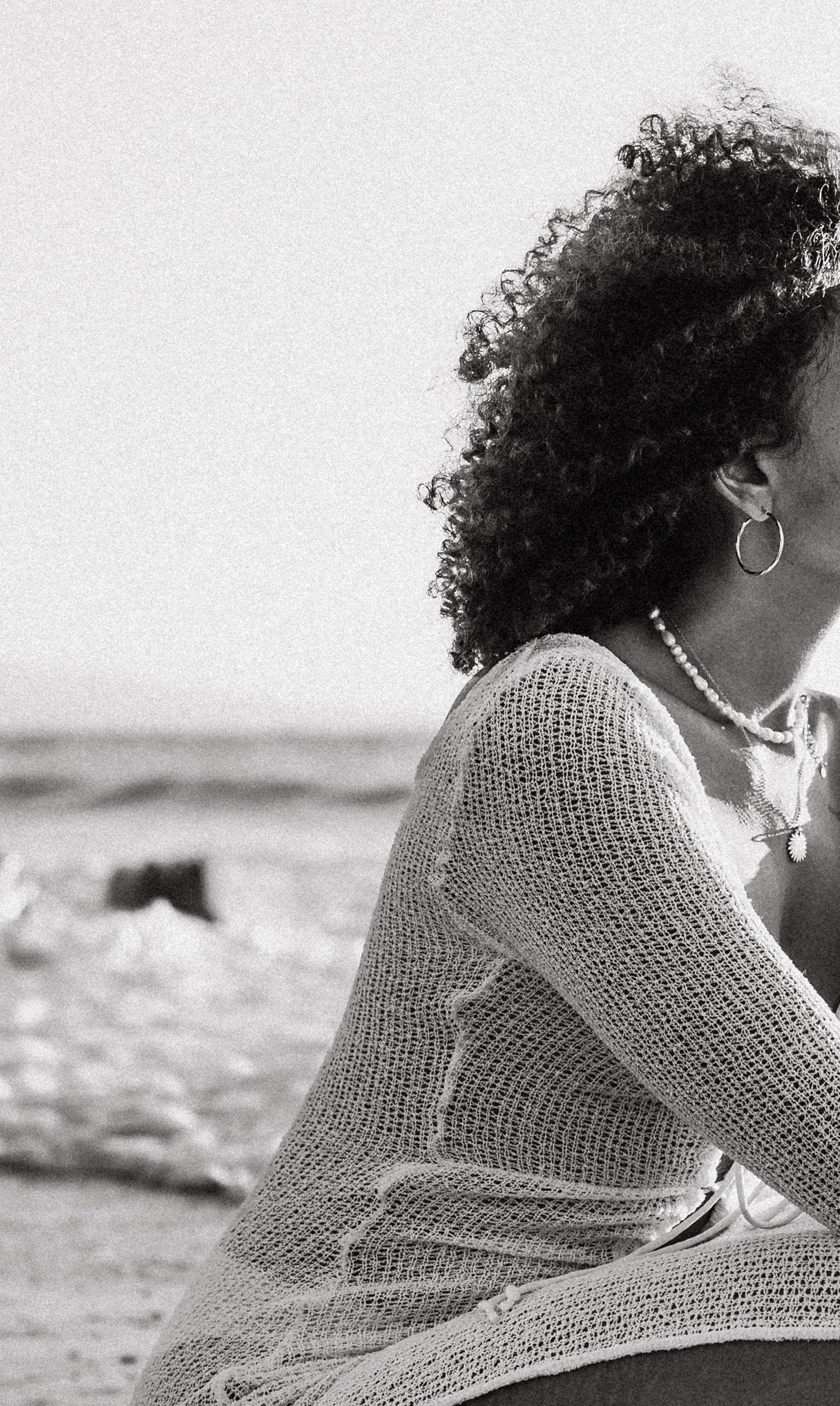 Une femme assise sur la plage avec des cheveux bouclés noirs portant un pull en maille, des boucles d'oreilles en anneau et un collier, en noir et blanc.
