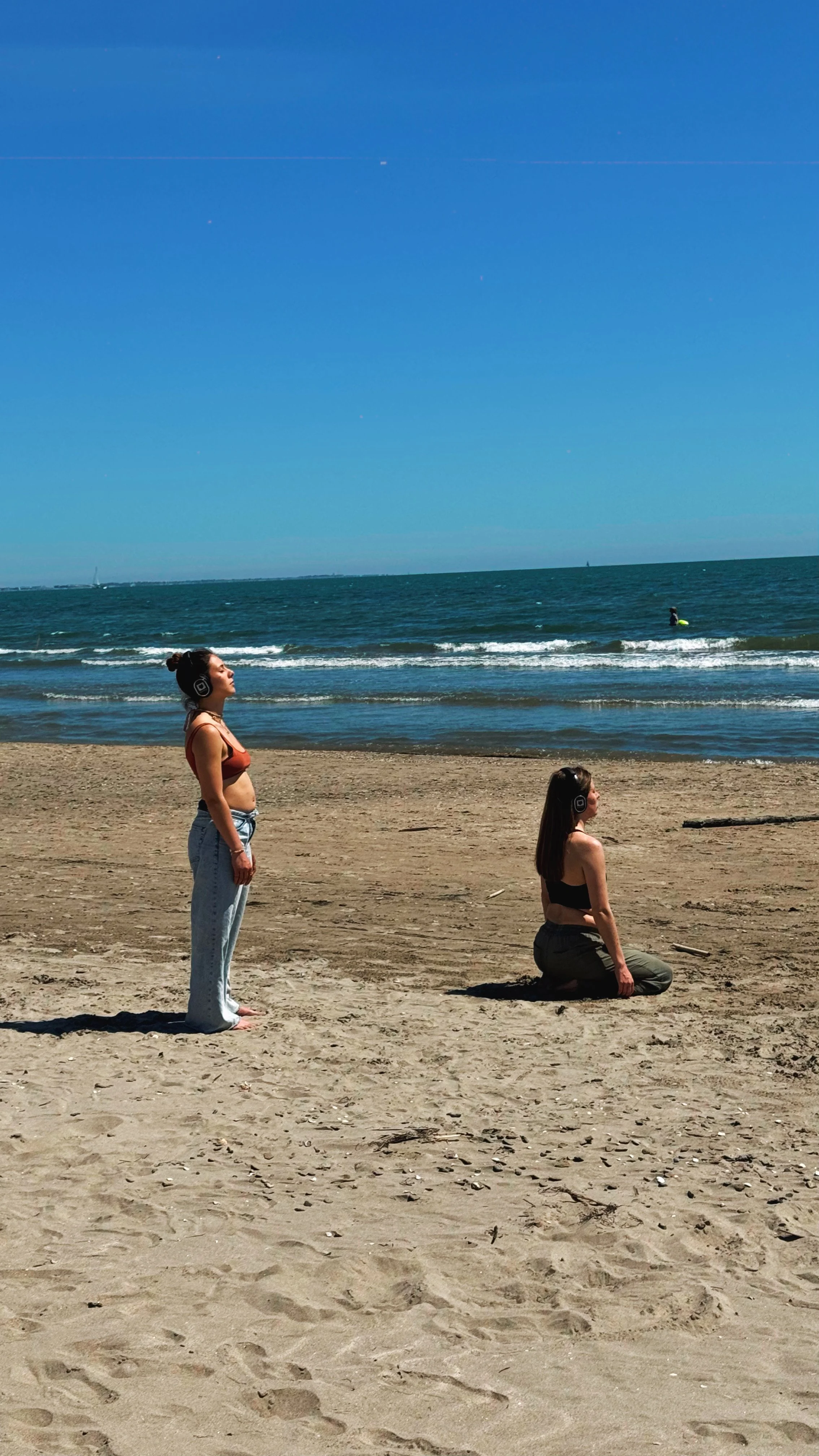 Deux femmes portant des écouteurs sur la plage, l'une debout et l'autre à genoux, face à la mer.