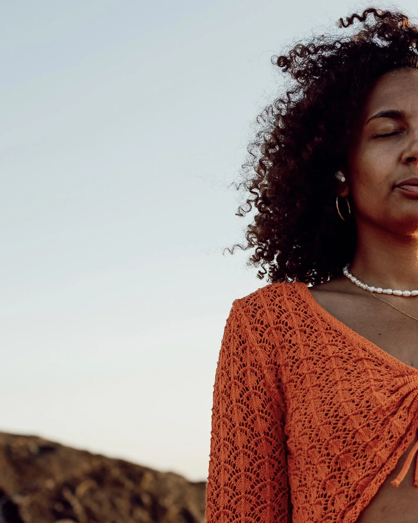 Femme africaine aux cheveux bouclés, portant un chemisier en crochet orange, des boucles d'oreilles en or, un collier de perles et un collier fin, dans un paysage naturel au coucher du soleil.