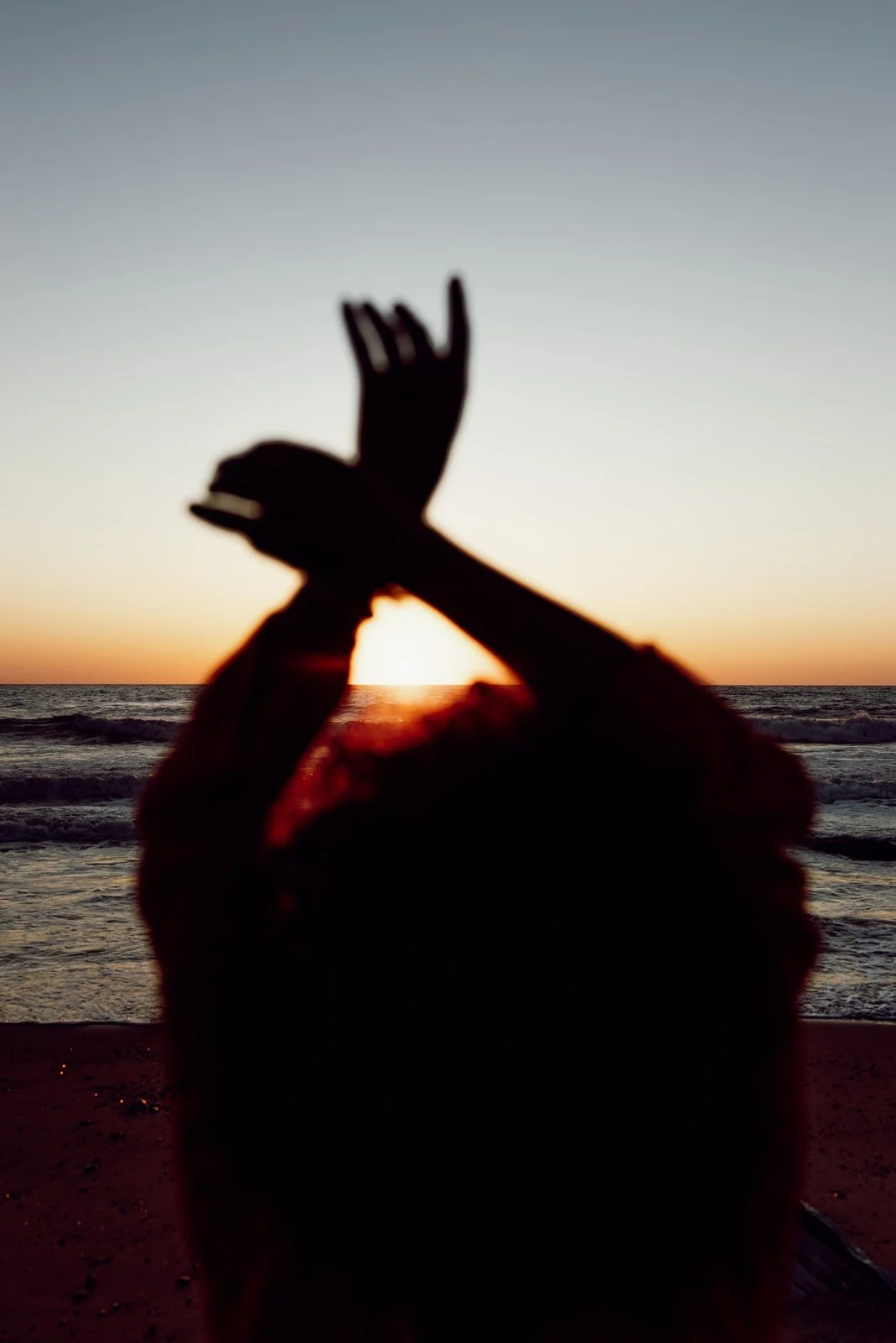 Silhouette d'une personne faisant un geste de paix avec une main, à la plage au coucher du soleil.