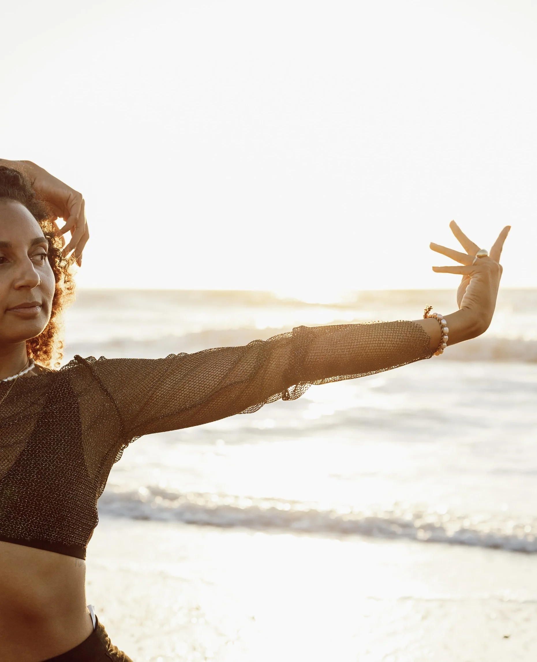 Une femme souriante au coucher du soleil sur la plage, portant un haut noir en filet et un bracelet, avec un bras étendu vers l'avant