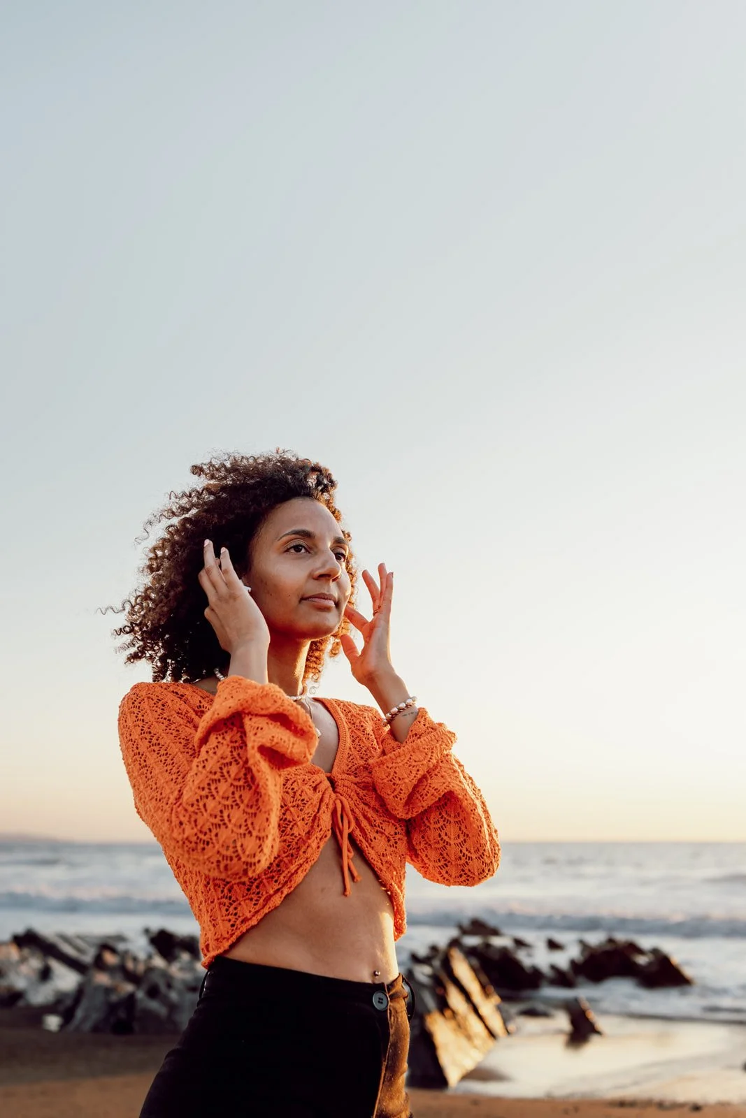 Jeune femme avec cheveux bouclés, portant un haut orange crocheté, se tenant sur la plage au coucher du soleil.