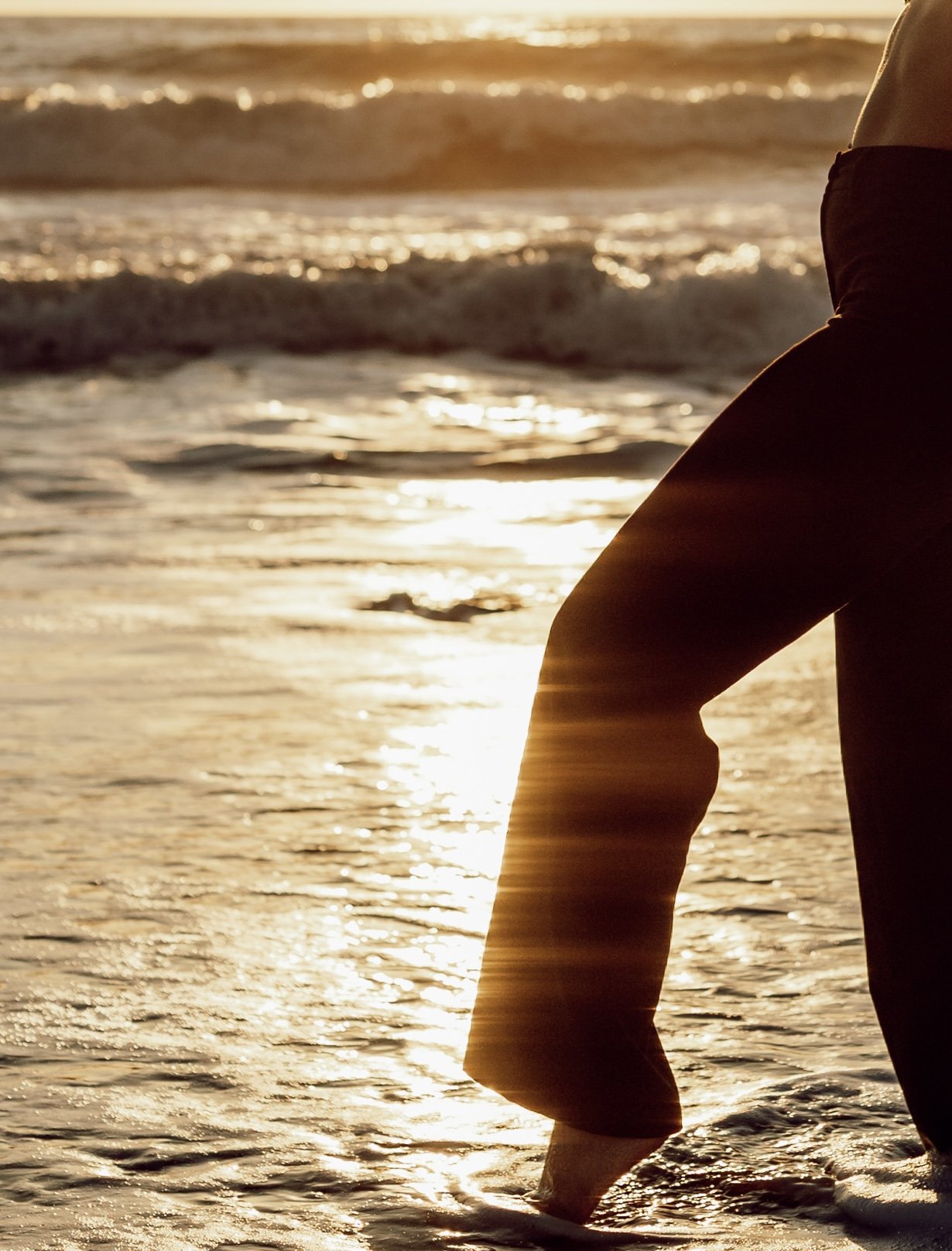 Une personne debout dans la mer au coucher du soleil, portant un pantalon noir, avec l'eau qui reflète la lumière dorée.
