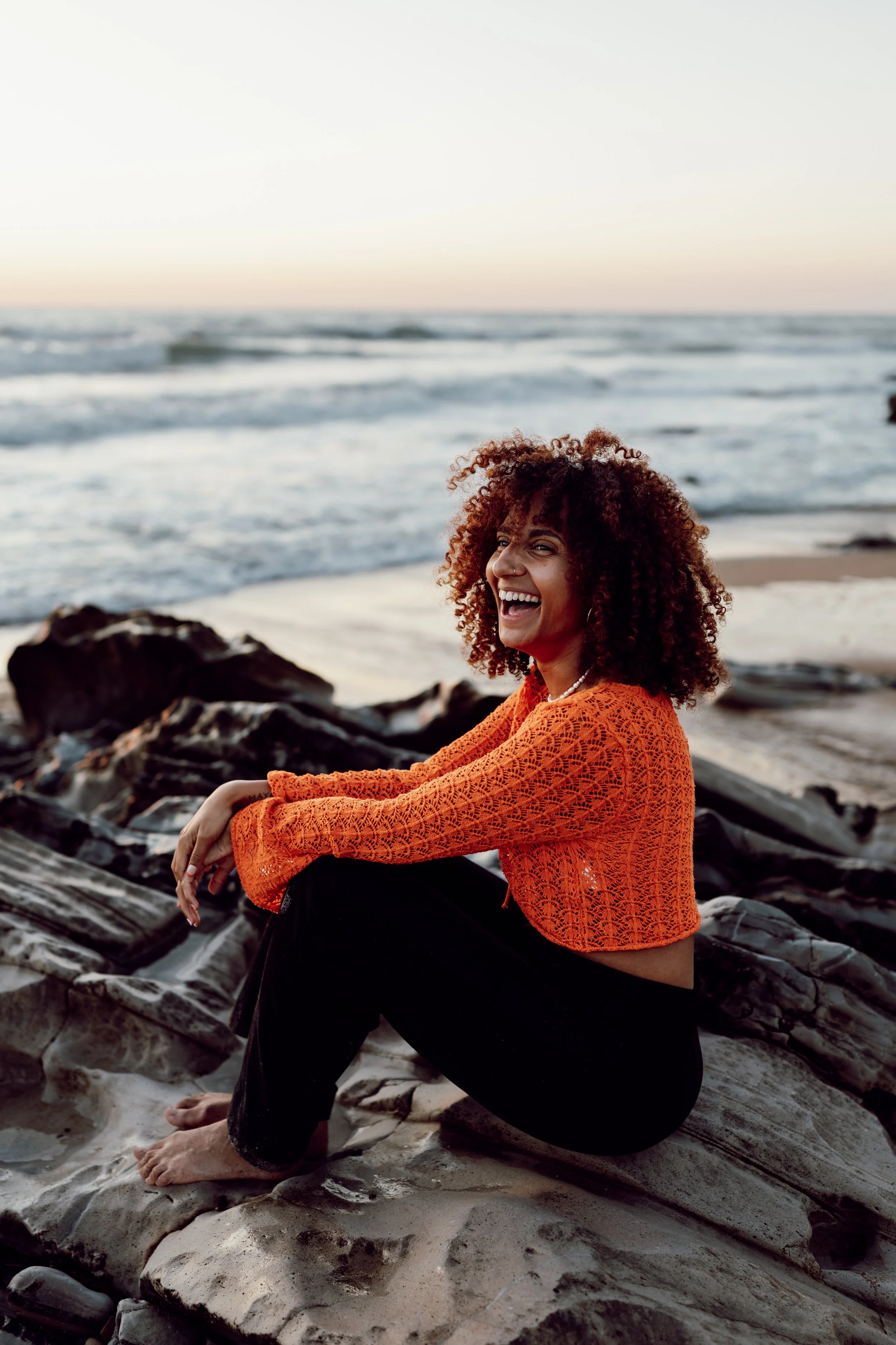 Une femme assise sur des rochers de plage, souriant, portant un pull orange en crochet, face à la mer au coucher du soleil.