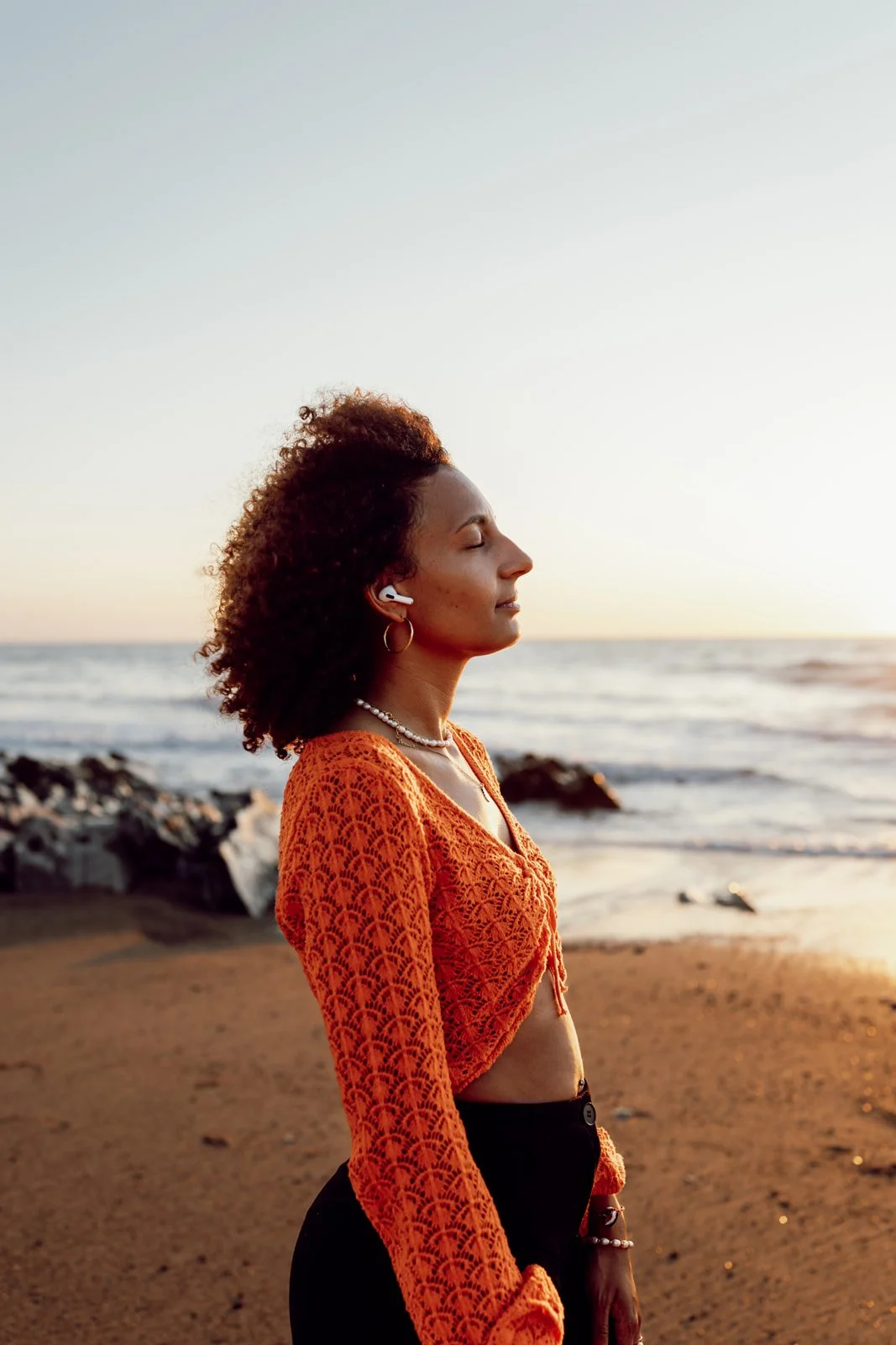 Une femme avec des cheveux bouclés, portant un haut en crochet orange et des accessoires, se tient sur la plage au coucher du soleil avec la mer en arrière-plan.