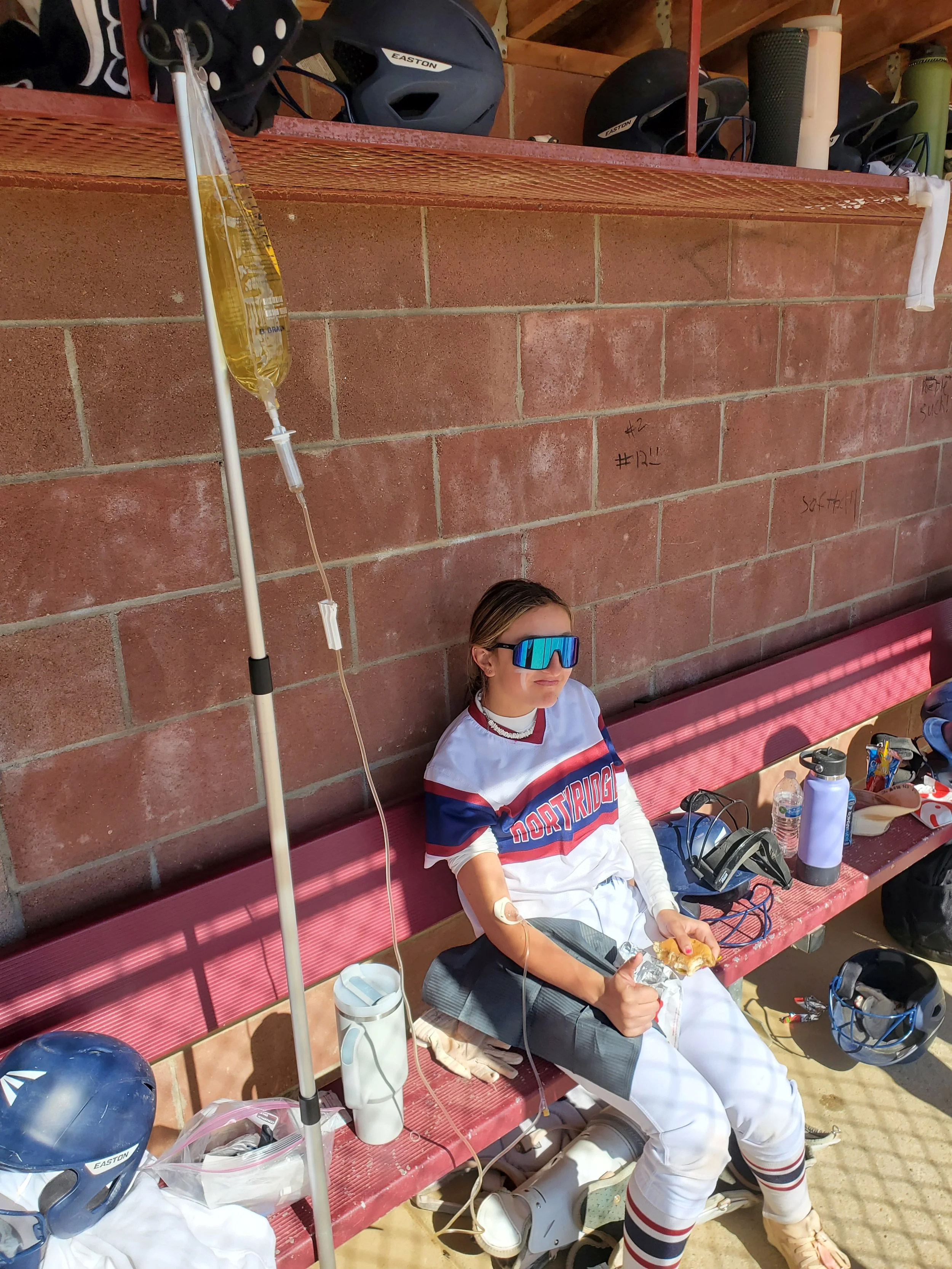 A young female baseball player sitting on a bench with an IV drip attached, wearing sunglasses, a baseball uniform, and holding a snack, with helmets and water bottles nearby.
