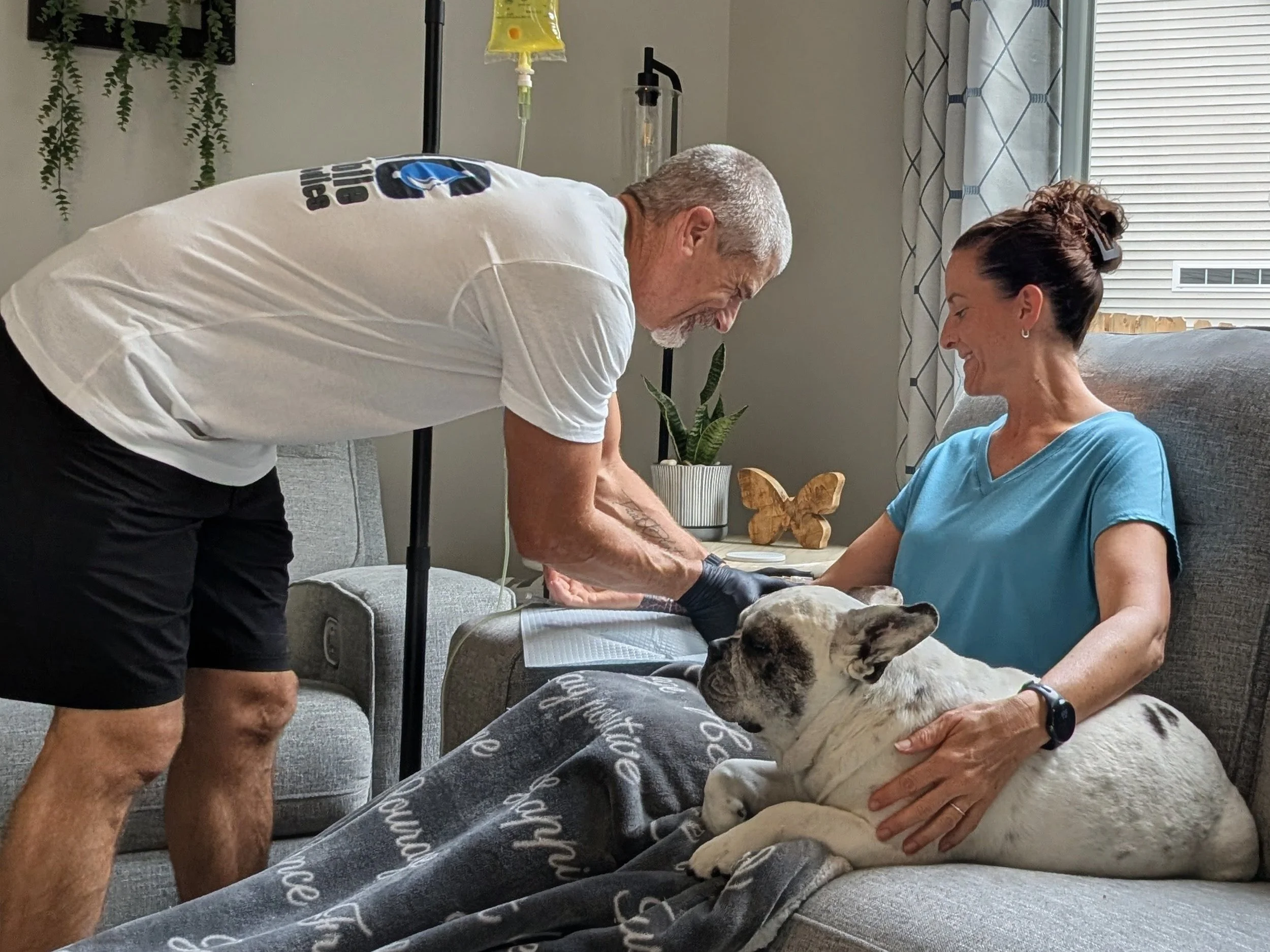 A woman sitting on a gray sofa with a bulldog lying on her lap, while an older man in a white T-shirt and black shorts leans over and interacts with her and the dog in a living room.