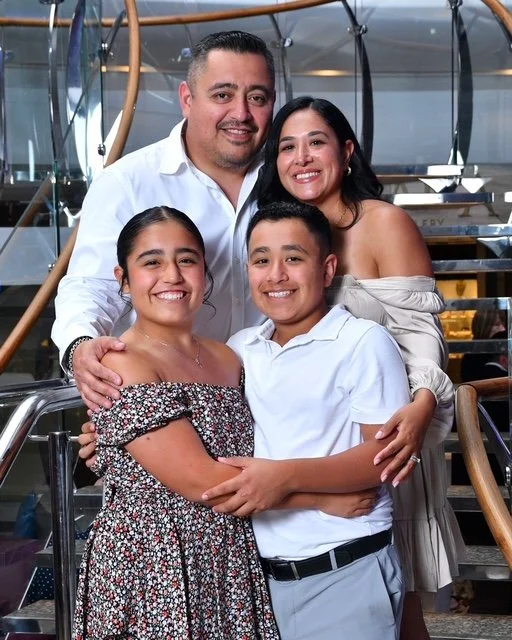 A family of four smiling and posing together on a cruise ship staircase. The father is wearing a white shirt, the mother in an off-shoulder beige dress, the daughter in a floral dress, and the son in a white shirt and gray pants.