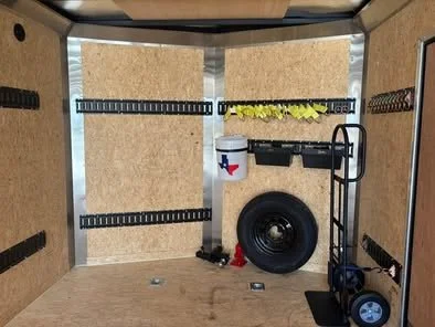 Empty walls with storage hooks, a large black tire, a wheelbarrow, and some hanging plants inside a small, unfinished garage or shed.
